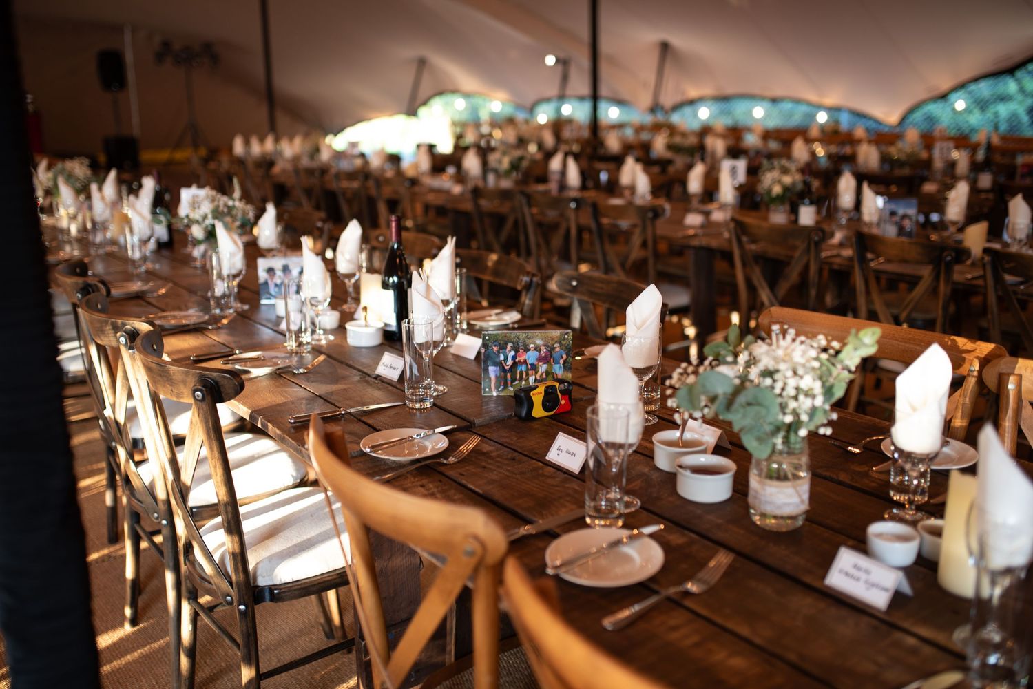 Rustic wooden tables set with white napkins and glassware inside a large wedding tent.