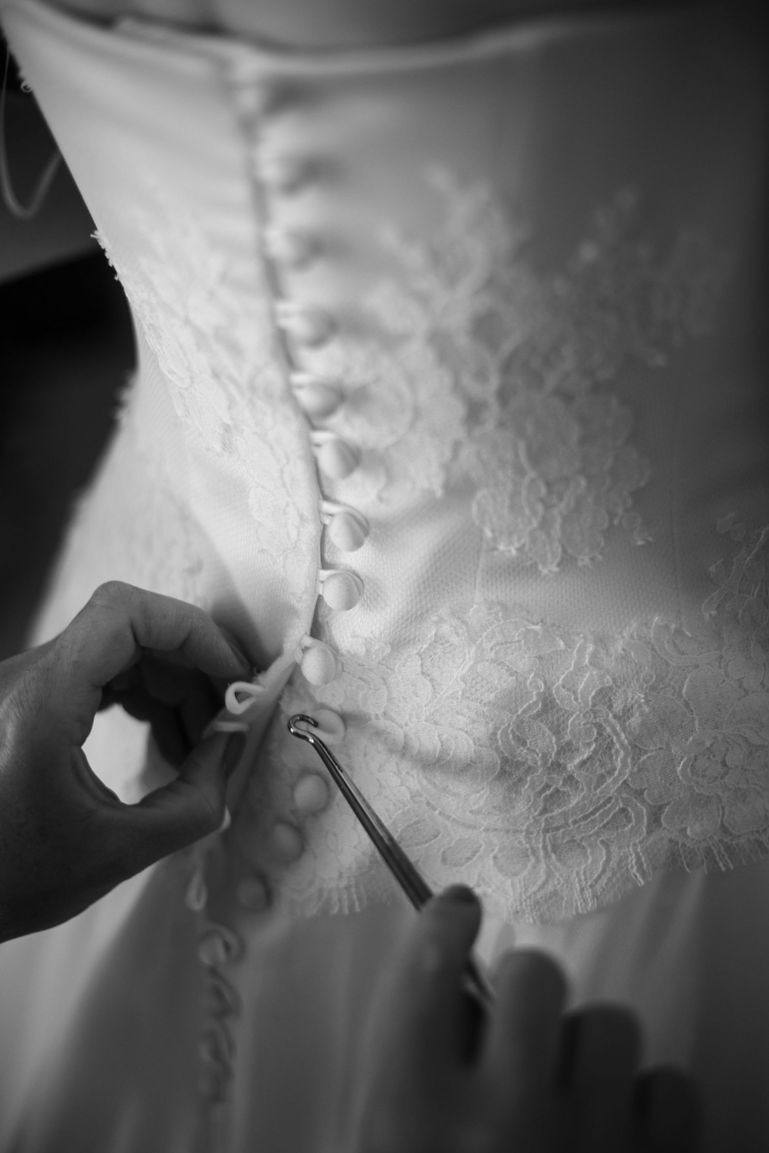 Black and white close-up of hands fastening buttons on the back of a lace wedding dress.