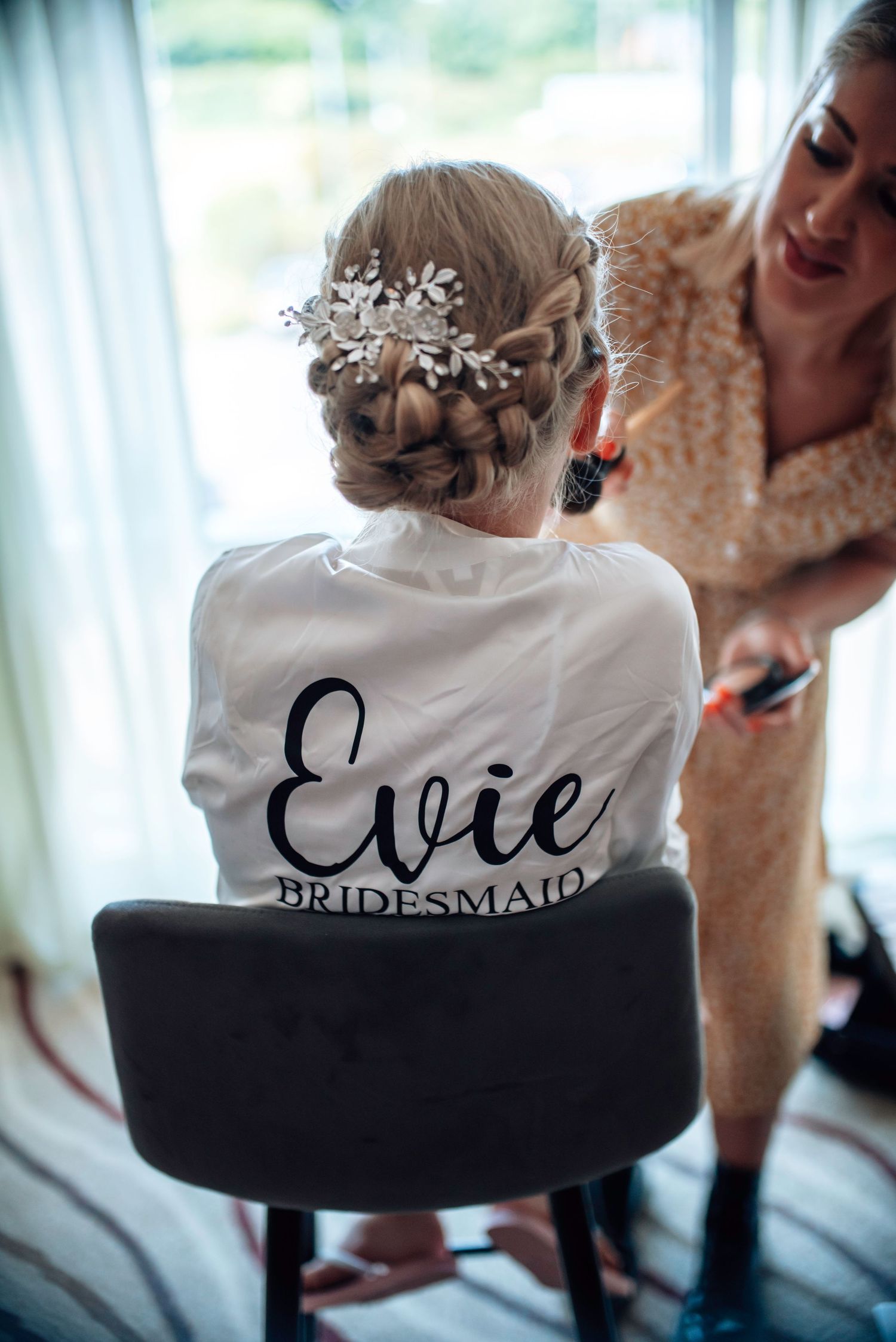 Bridesmaid Evie in personalized white robe with elegant braided updo adorned with floral hairpiece.