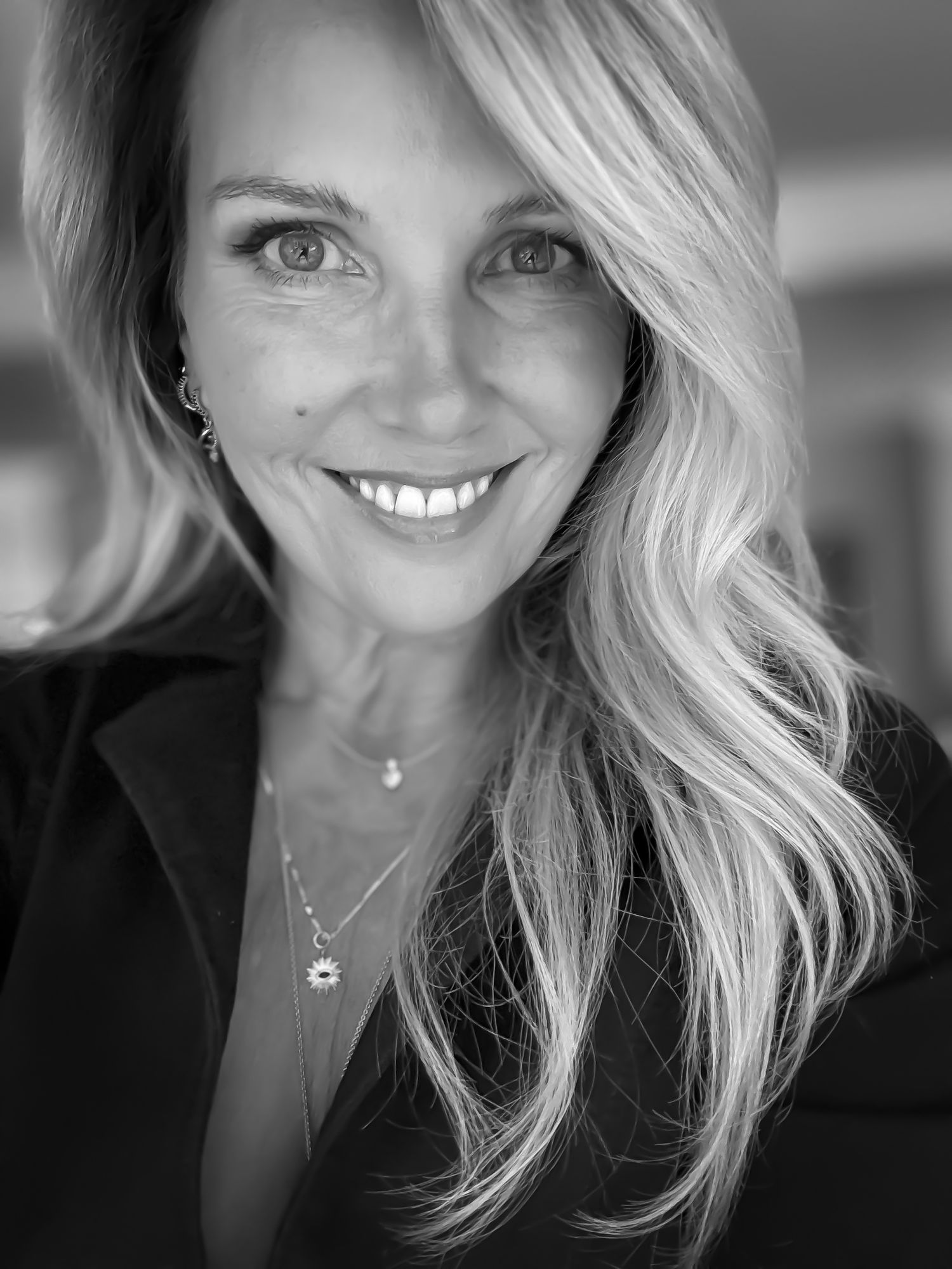Black and white close-up portrait of smiling blonde woman with layered necklaces. Heidi Barroll Brown