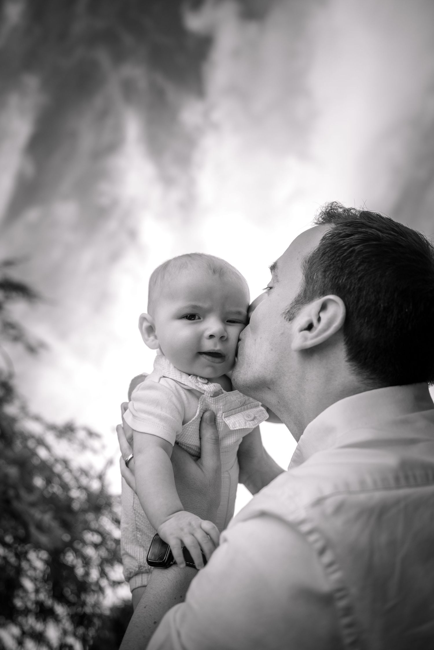 Black and white portrait of father kissing his smiling baby outdoors.
