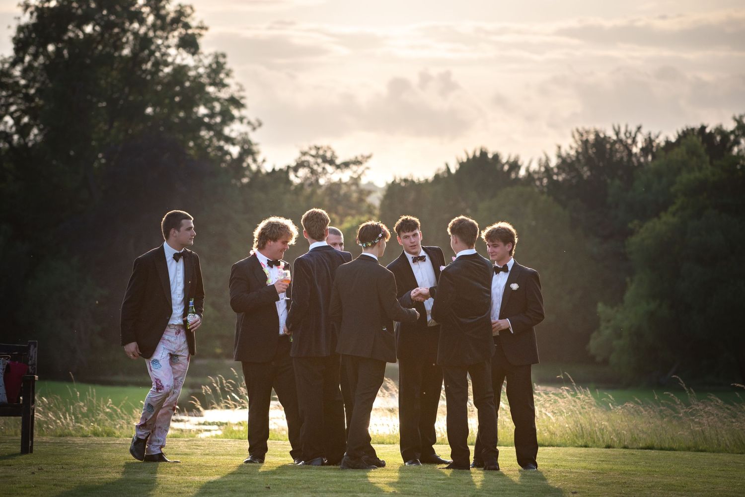 Group of men in black tuxedos socializing on a lawn at sunset.