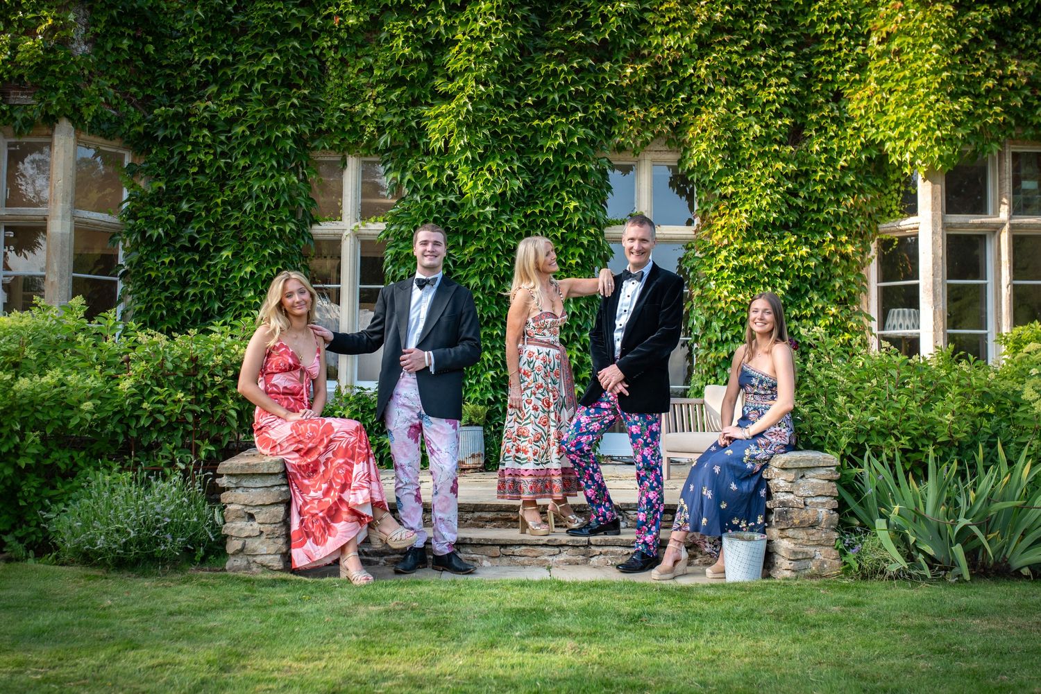 Group of five elegantly dressed wedding guests posing on stone steps in front of an ivy-covered manor house.