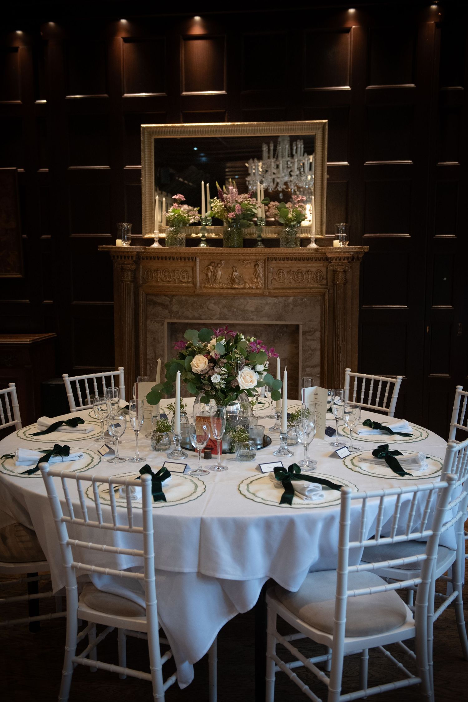 Elegant wedding reception table set with white linens, dark green ribbon napkins, and floral centerpiece near ornate fireplace.
