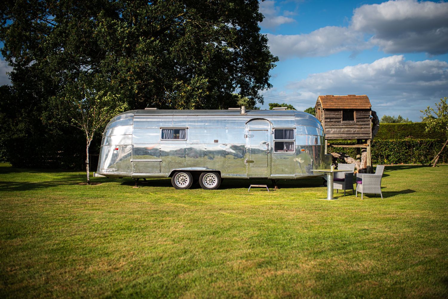 Shiny vintage Airstream trailer parked on green grass beside a wooden treehouse under blue skies.