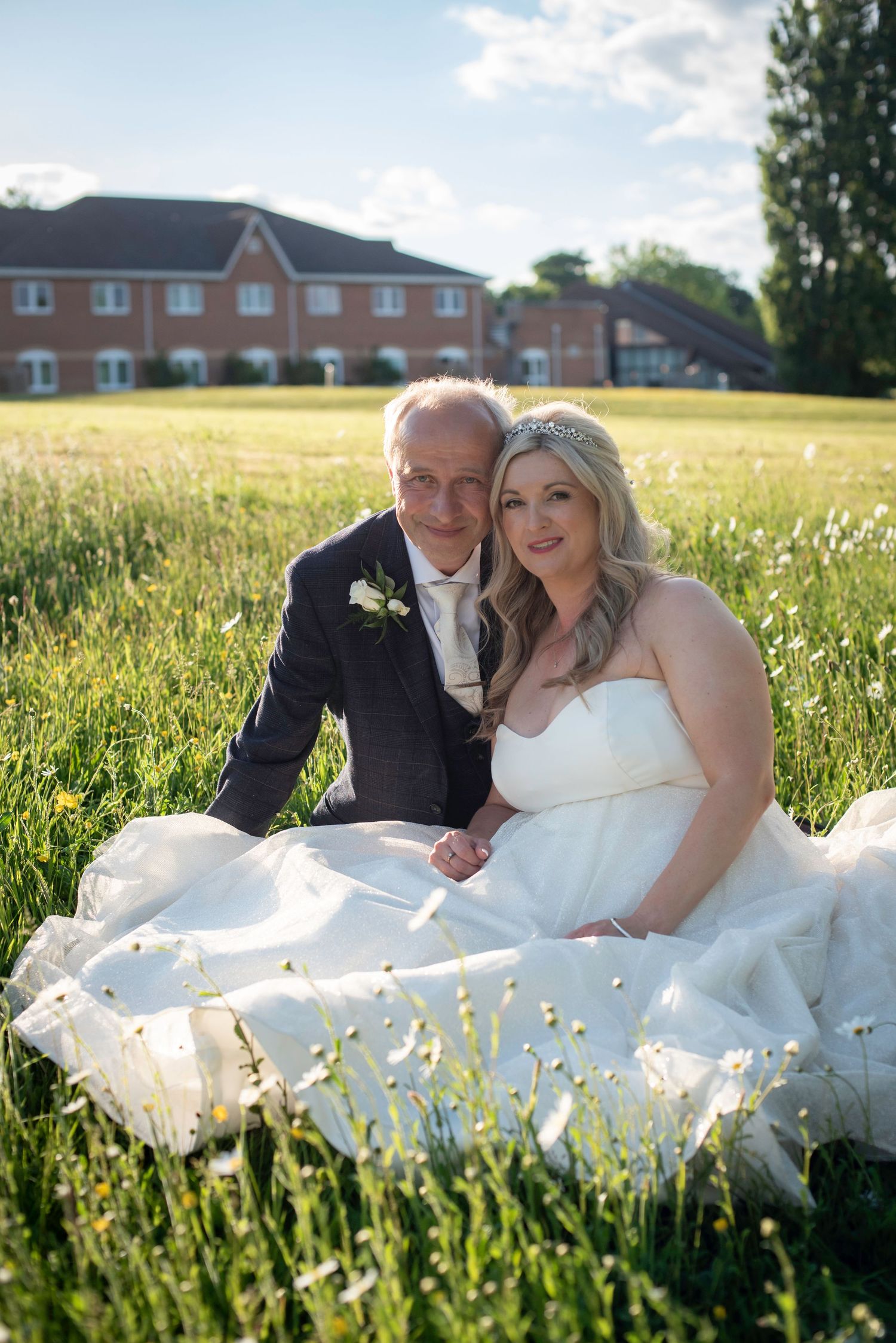 Bride and groom sitting in a field of daisies with a brick building in the background.