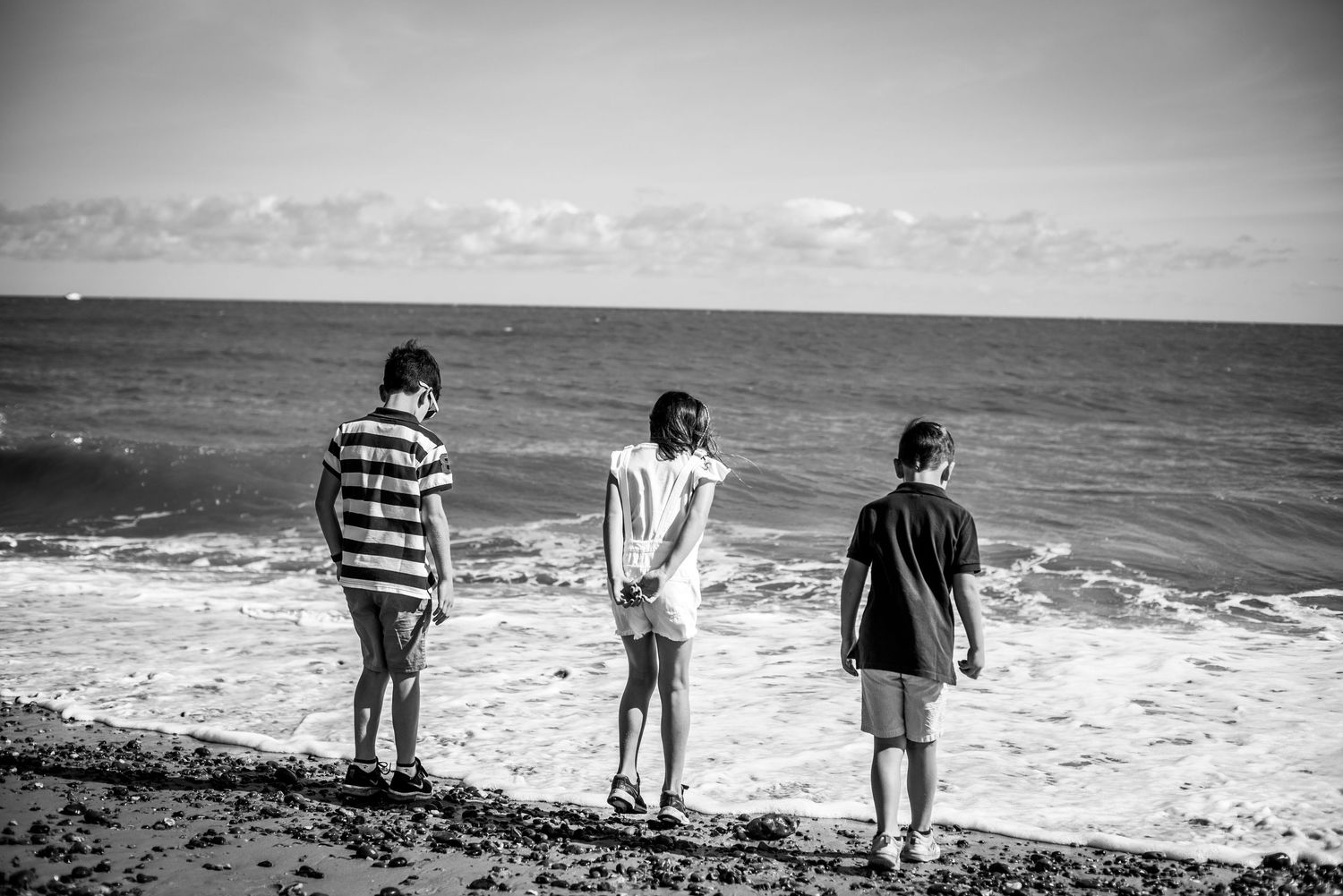 Black and white photo of three boys standing on rocky beach looking out at ocean waves.