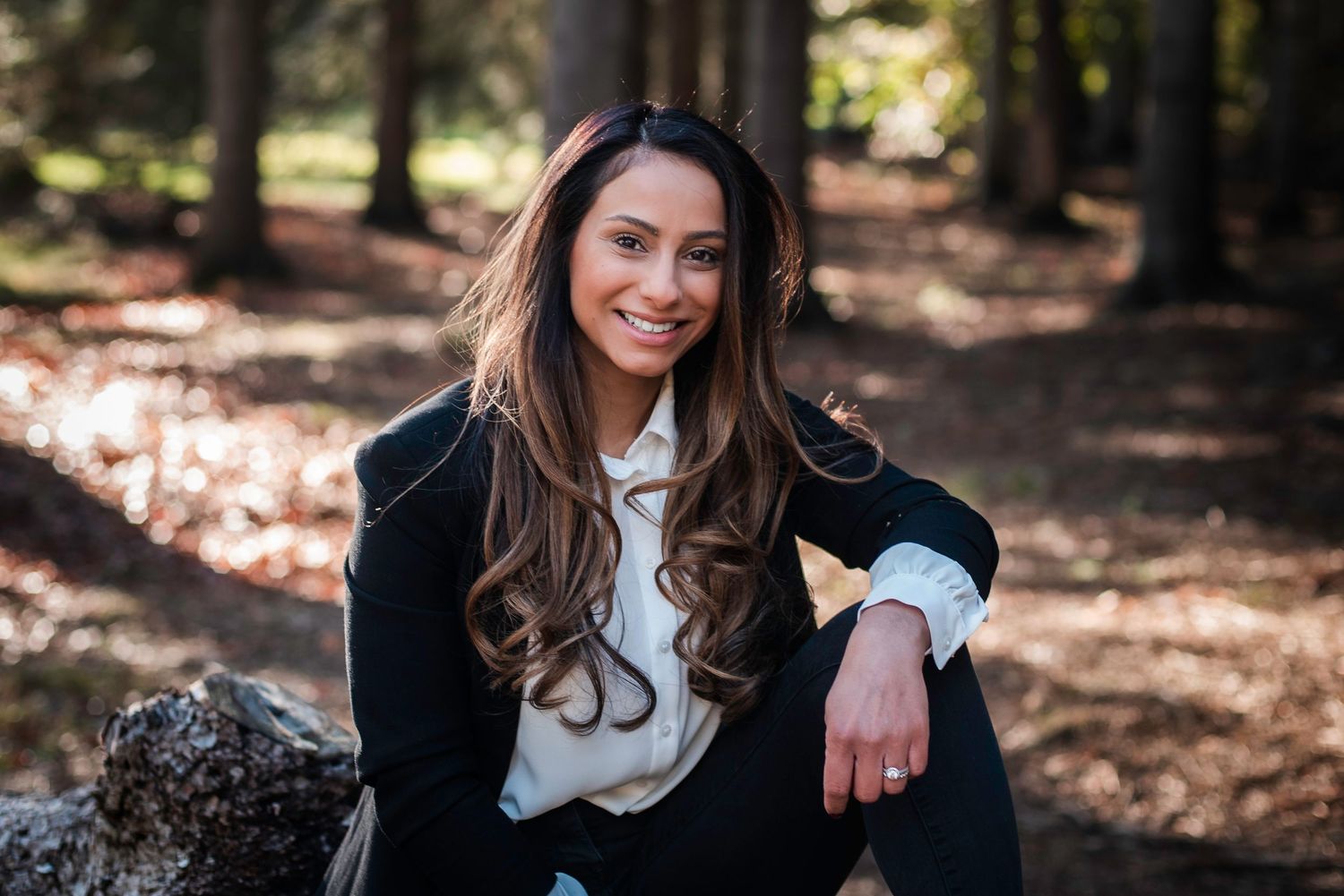 Young brunette woman smiling warmly while sitting on rocks in a sunlit forest setting. business portrait