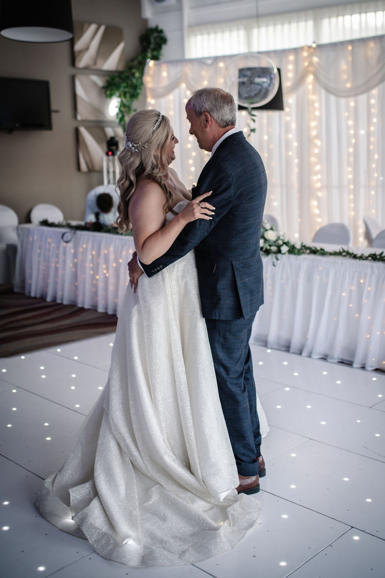 Bride and groom share their first dance on a white LED dance floor with fairy lights in the background.