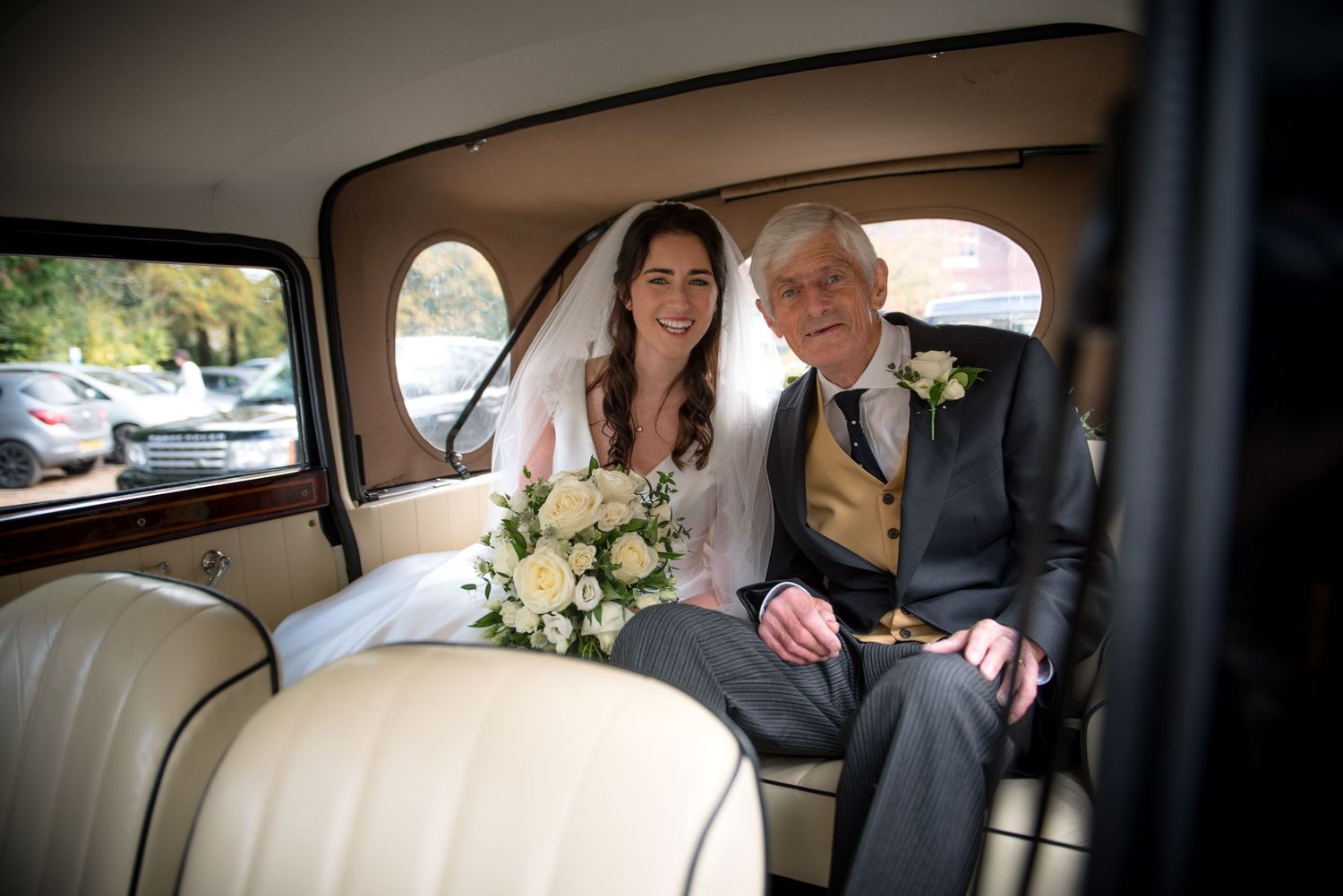 Bride holding bouquet seated beside her father in a classic vintage wedding car.