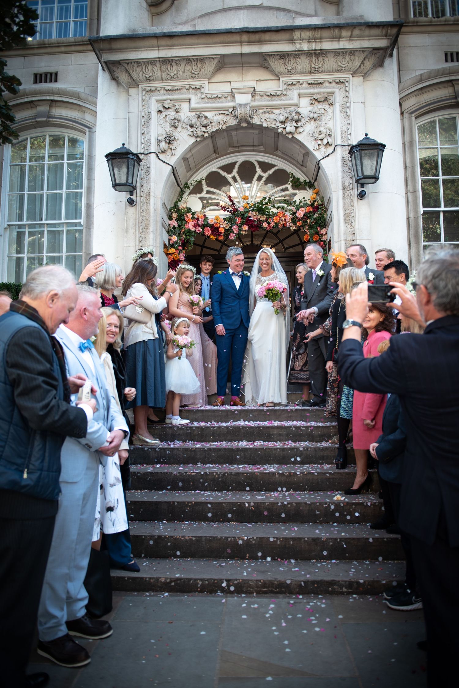 Newlyweds exit grand building steps as guests throw confetti with floral arch backdrop at Chelsea Town Hall Wedding.