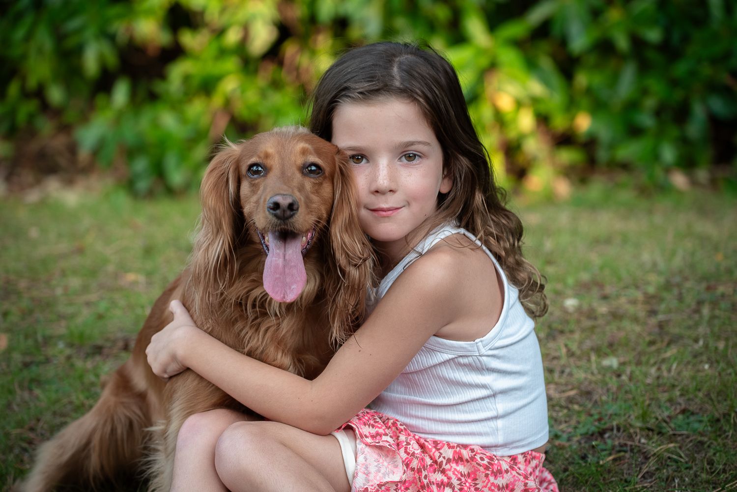 Young girl hugging golden retriever dog while sitting on grass outdoors.