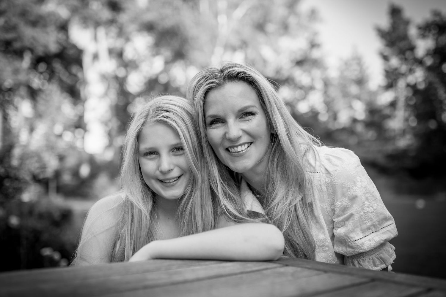 Black and white portrait of a smiling mother and daughter leaning on a wooden table outdoors.