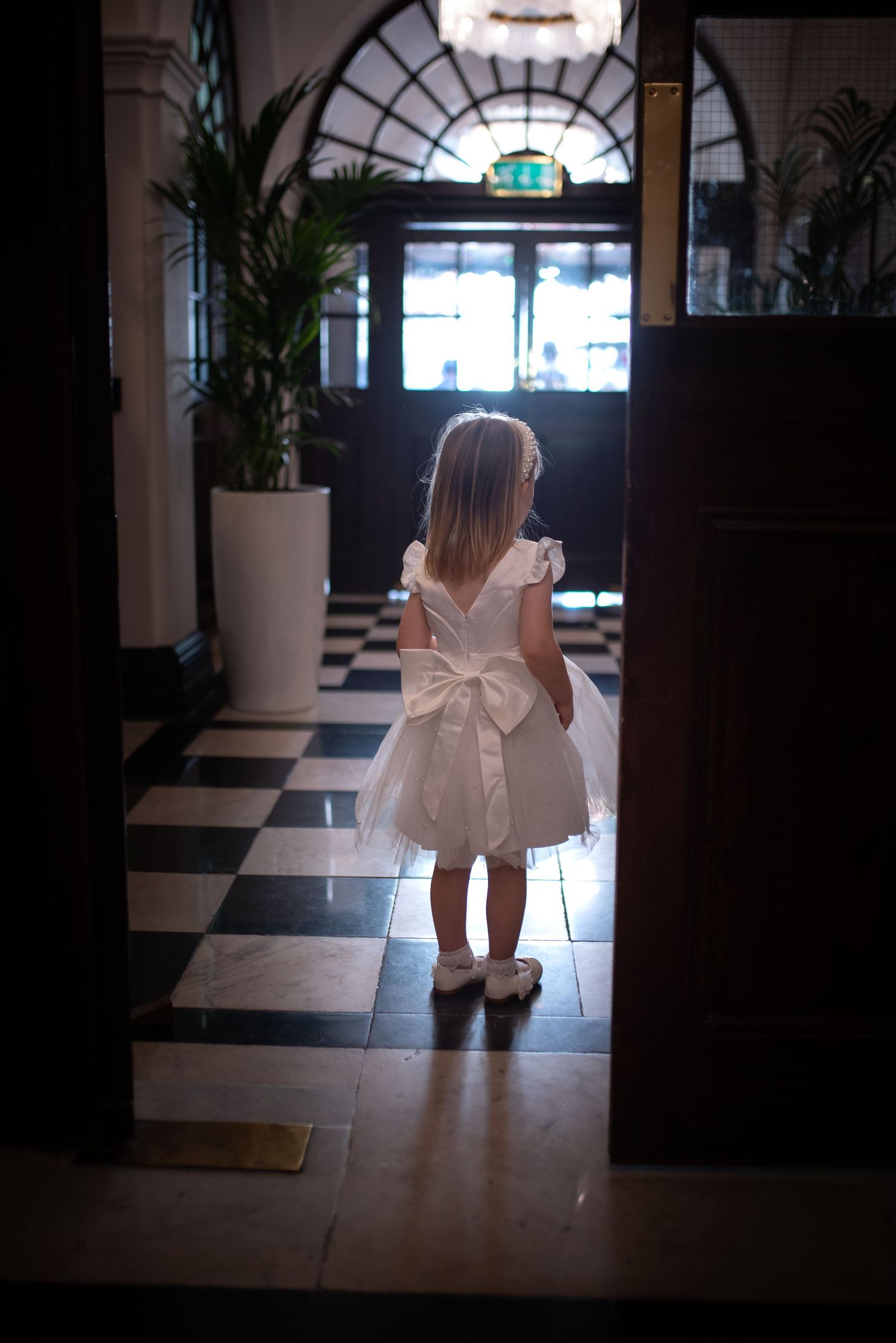 A little flower girl in a white dress with bow stands in an elegant checkered lobby doorway.