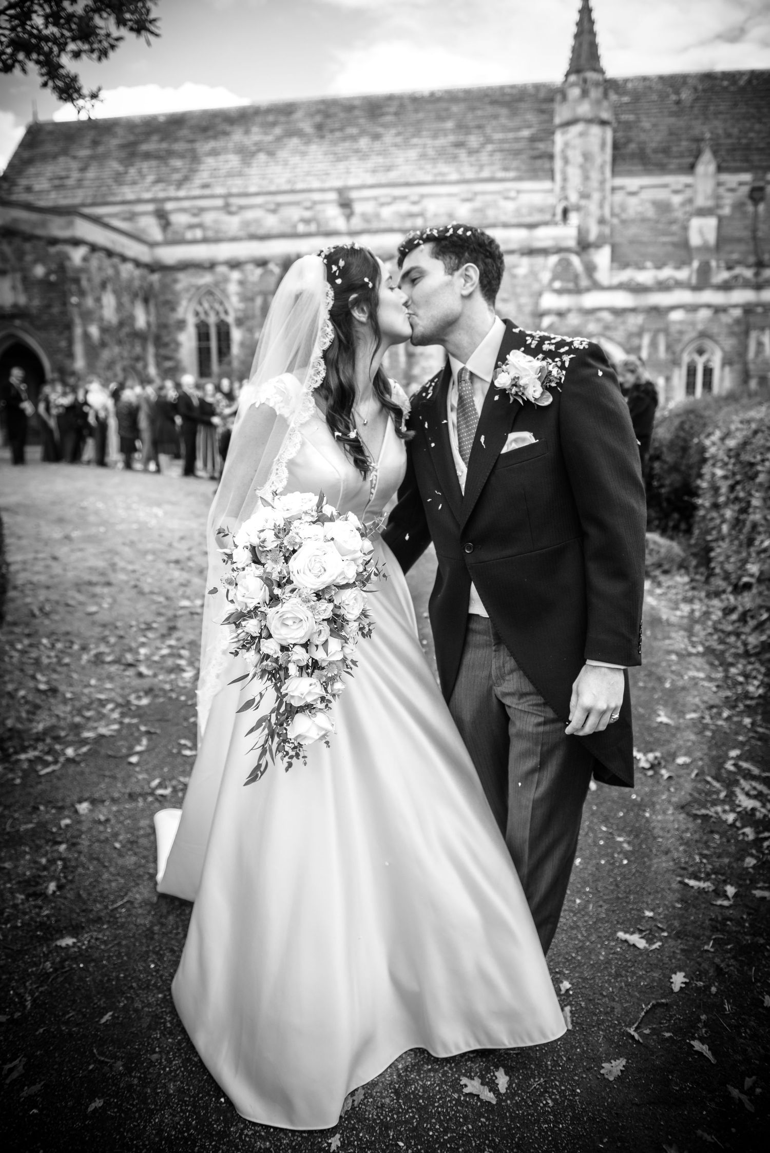 Black and white photo of a bride and groom sharing a kiss outside a historic stone church. St Saviours Church, Brockenhsurt.