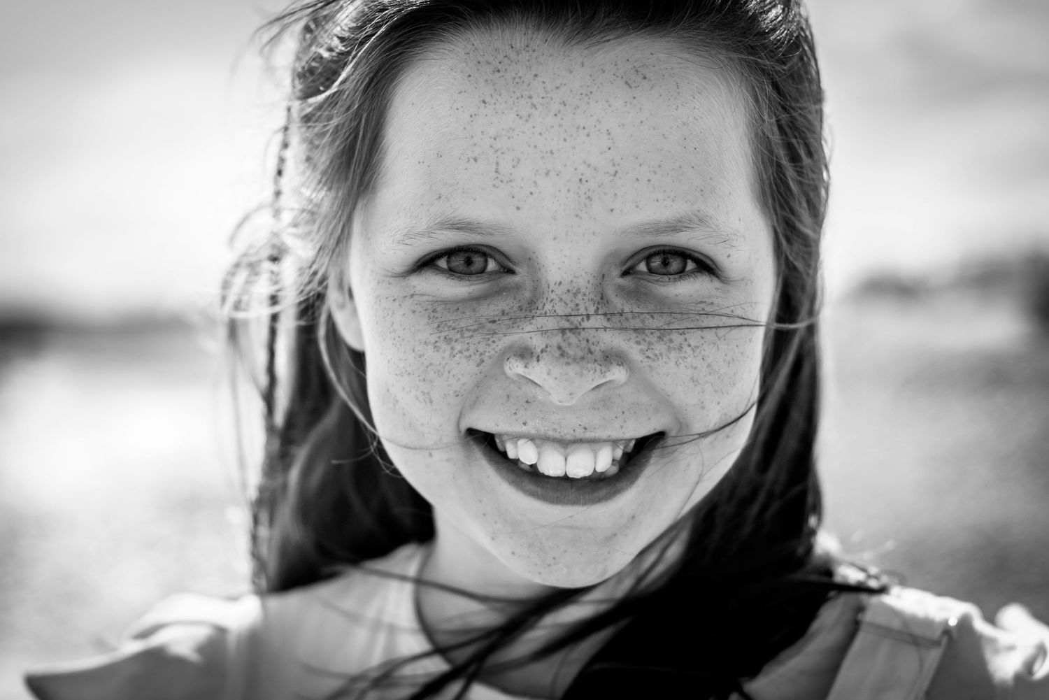 Black and white close-up portrait of smiling freckled girl with windswept hair outdoors.