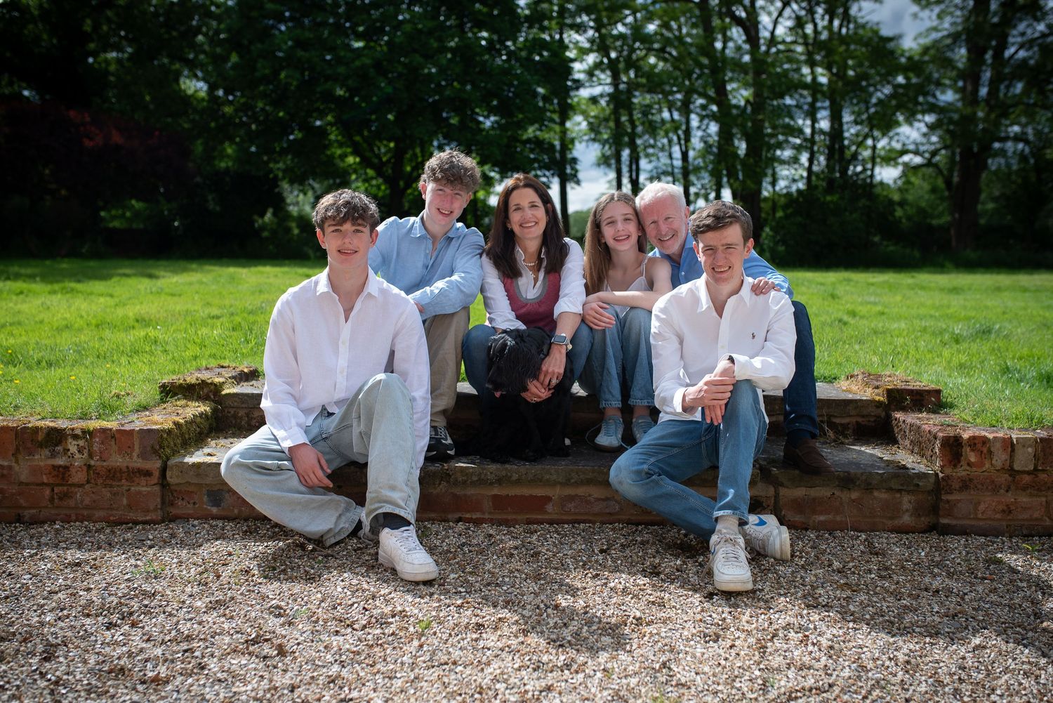 Family of six posing on stone steps surrounded by trees in a bright sunny outdoor setting.