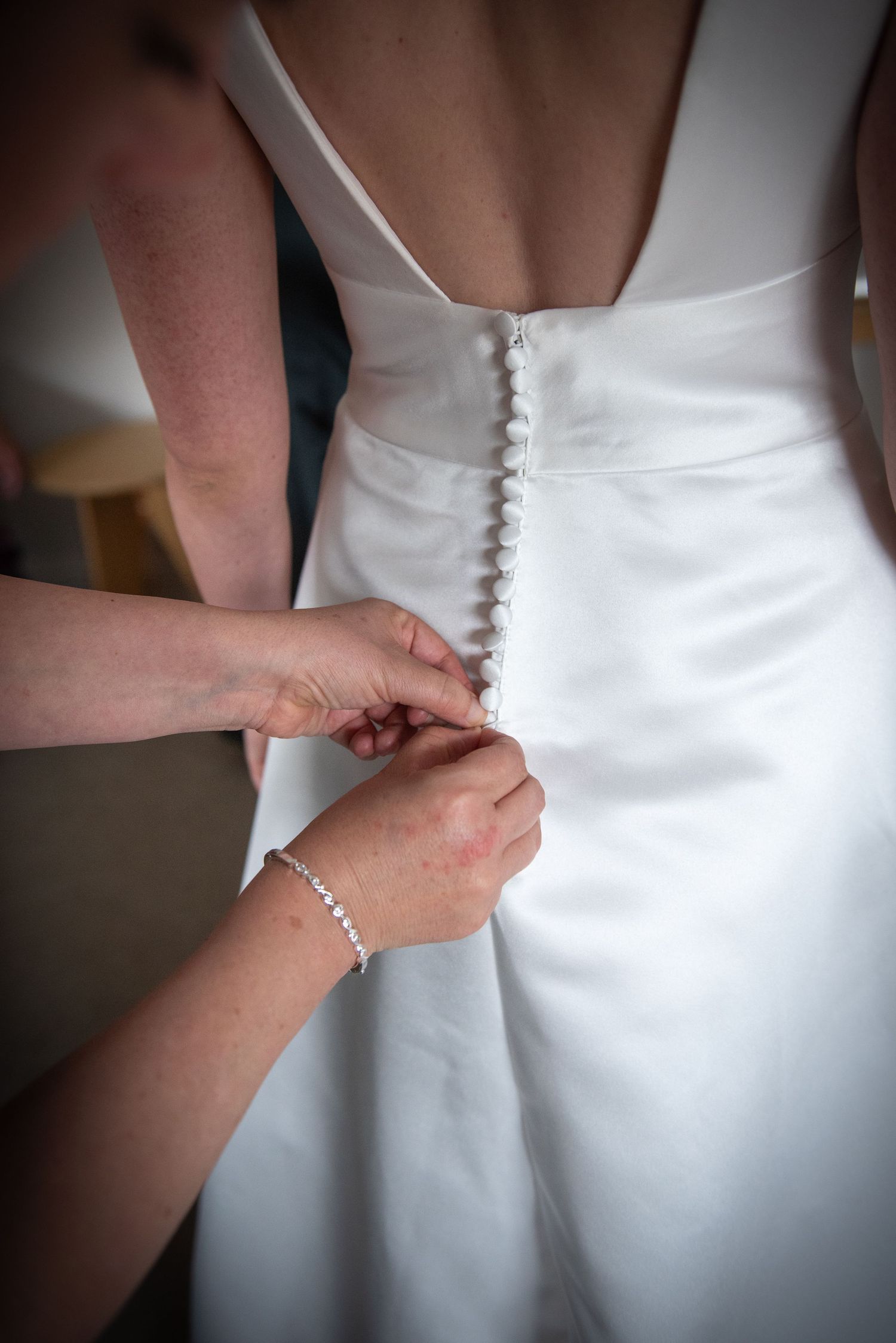 Hands fastening pearl buttons on the back of a white satin wedding dress.