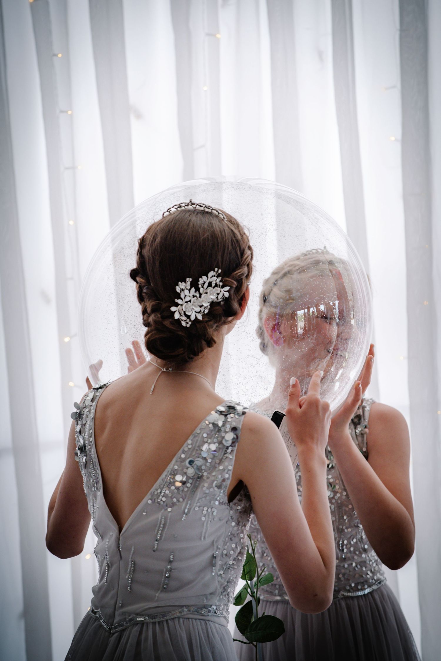 Two bridesmaids in silver sequin dresses hold large transparent balloons near white curtains.