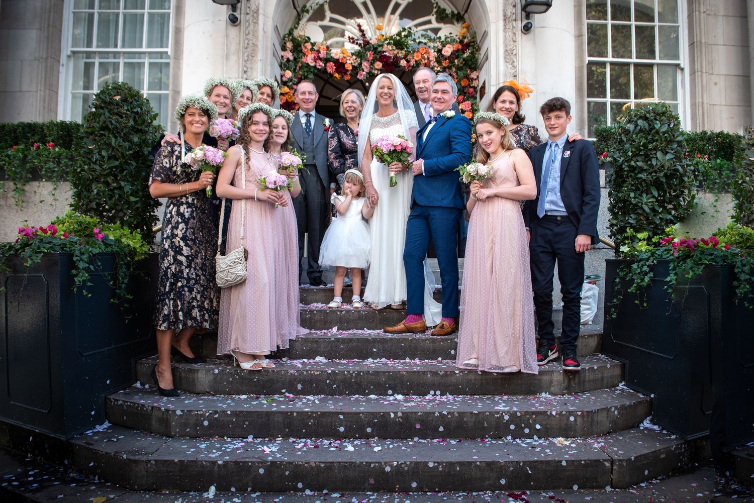 Wedding party group photo on steps with bridesmaids in pink dresses and floral arch backdrop. London Wedding at Chelsea Town Hall.