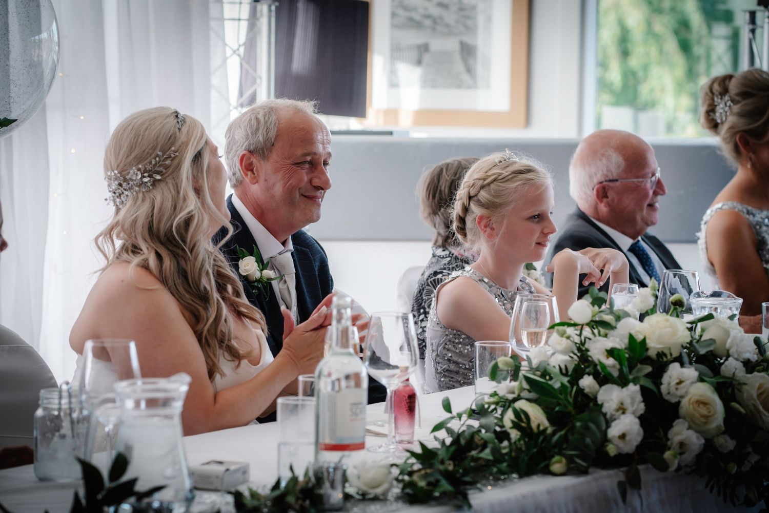 Wedding guests smile and laugh at elegantly decorated reception table with floral centerpiece.