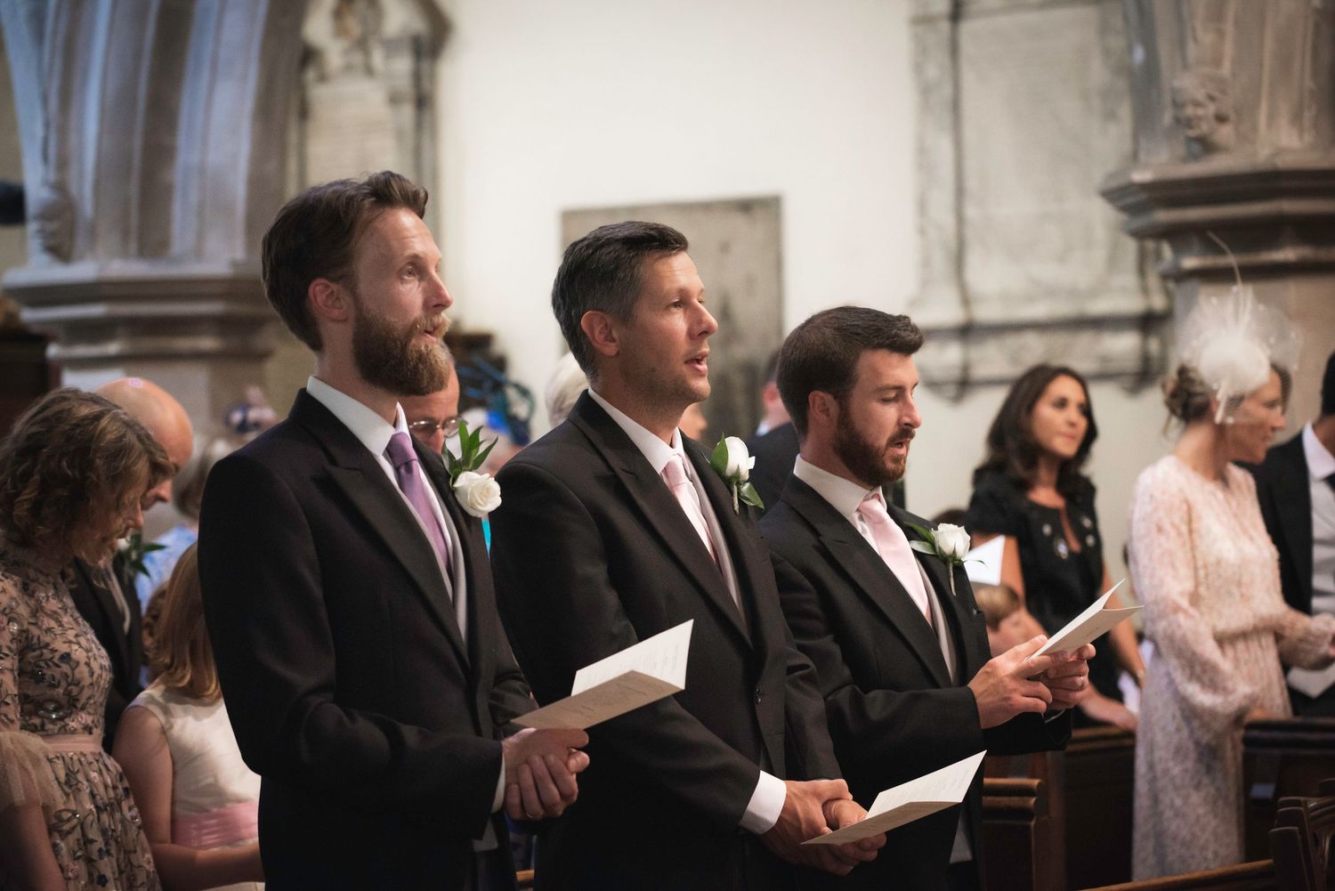 Three groomsmen in dark suits holding order of service booklets during church wedding ceremony.  St Michaels Church, Bray