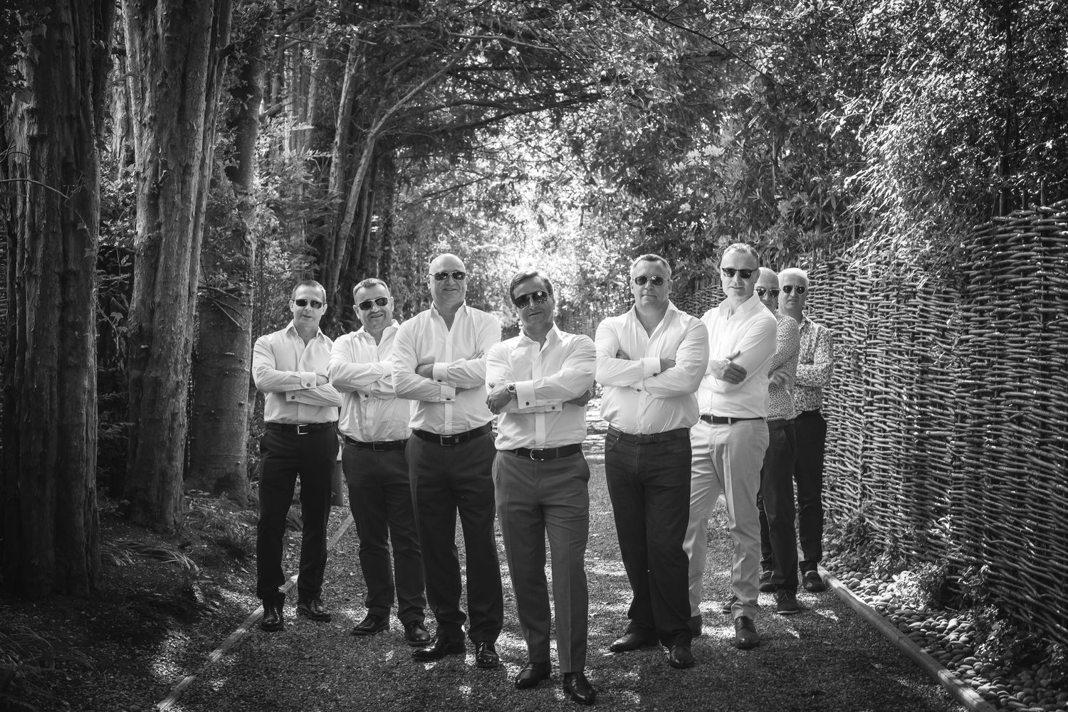 Group of groomsmen posing with arms crossed on a tree-lined path in black and white.