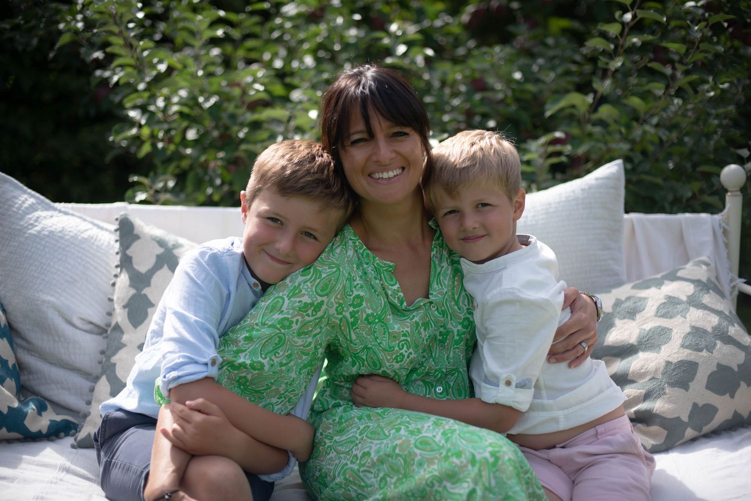 A smiling mother in a green floral dress hugs her two young sons on a garden bench. family portrait
