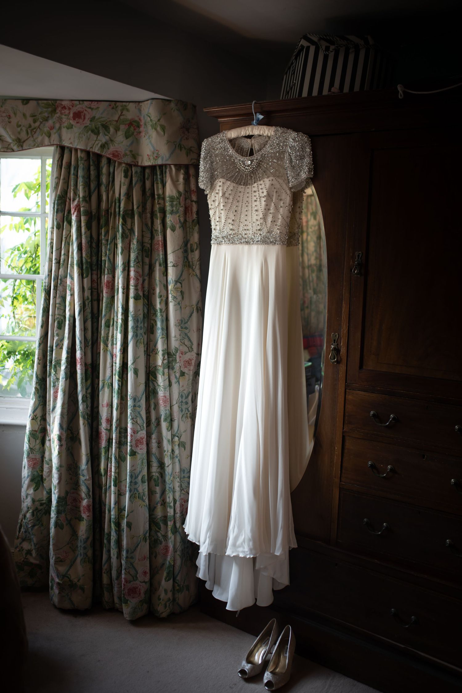 Beautiful beaded cap sleeve wedding gown hanging in a bedroom with floral curtains and heels.