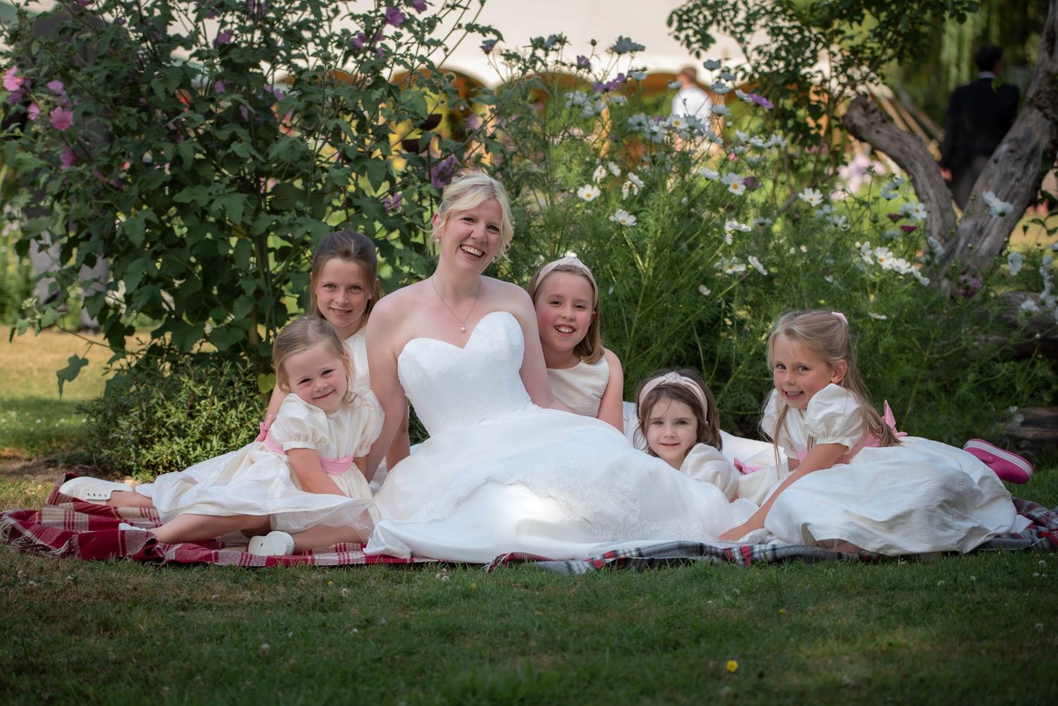 Bride in white gown sits on grass with five young flower girls in cream dresses at outdoor wedding.