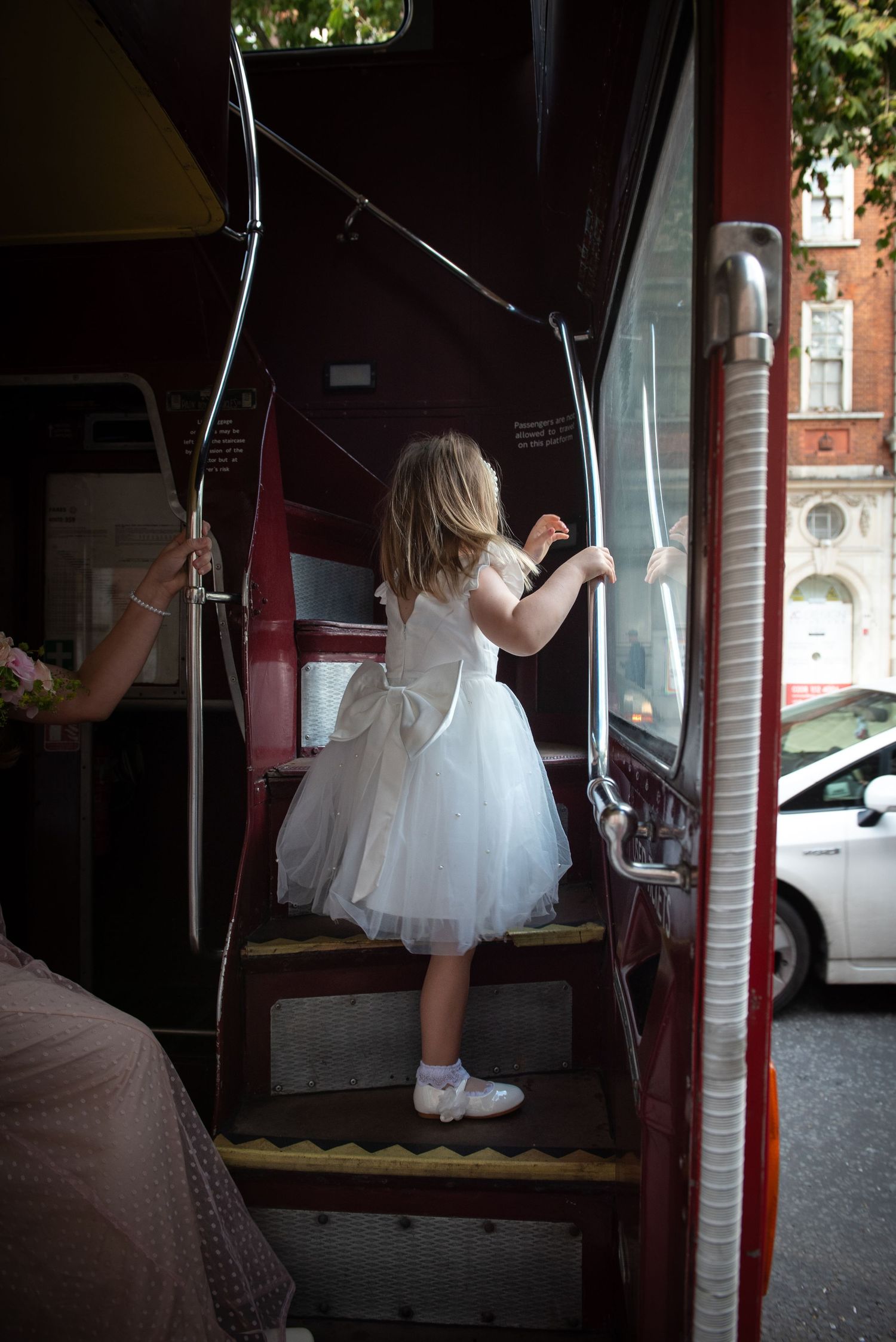 Young flower girl in white tulle dress boarding a red double-decker bus after wedding. Chelsea Town Hall Wedding