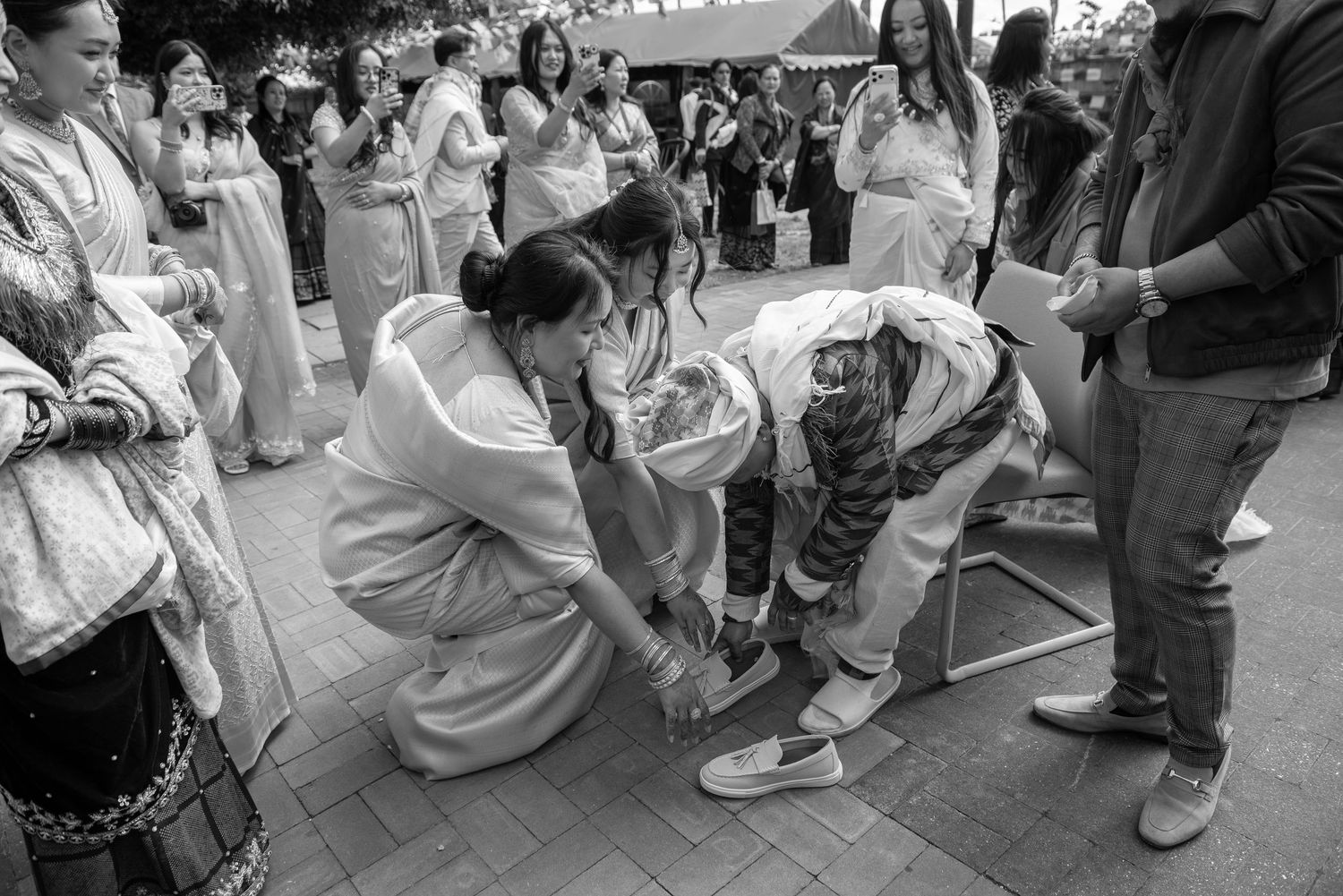Guests in traditional attire help tie shoes during a cultural wedding ceremony outdoors.