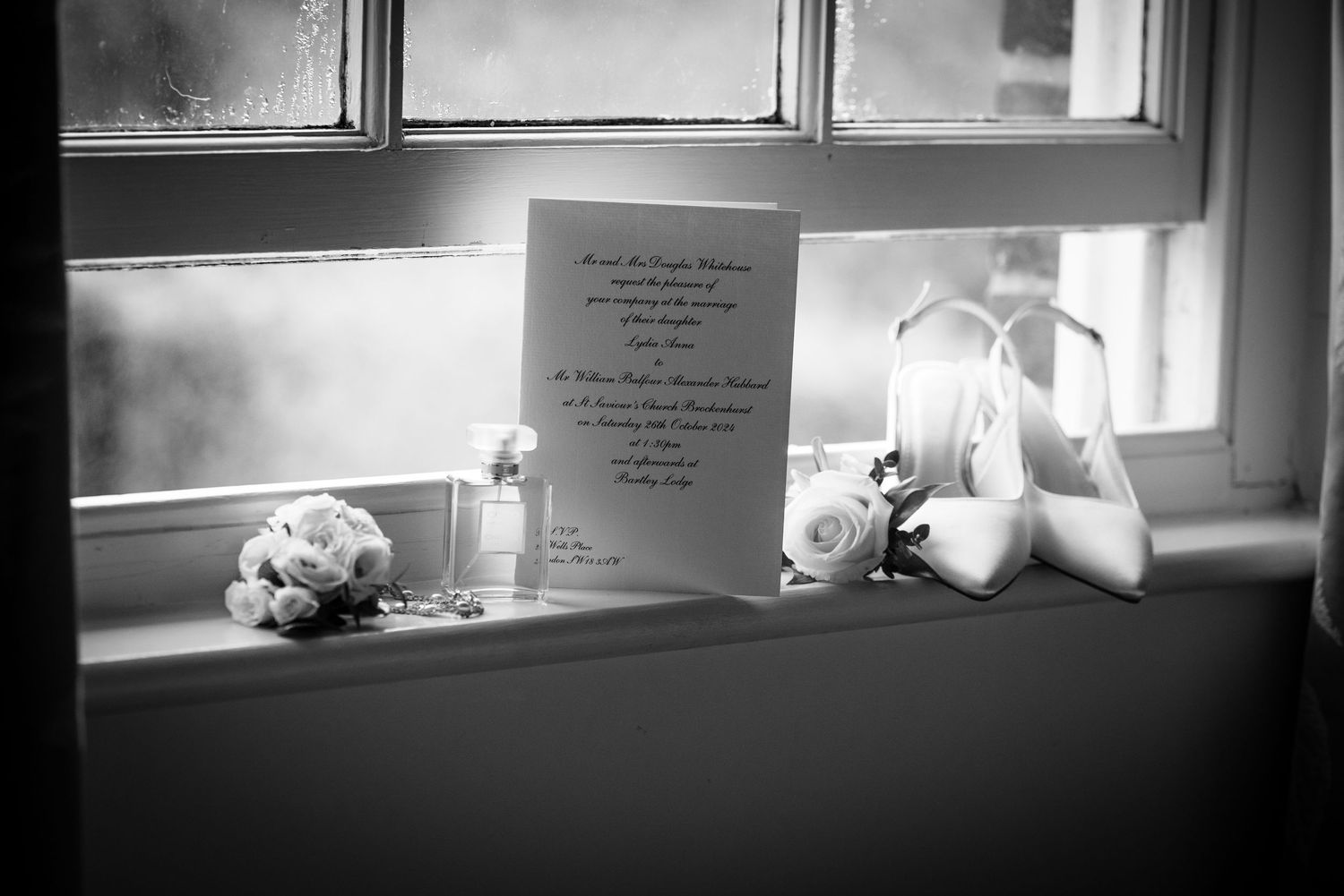 Wedding invitation, bridal shoes, and bouquet on a windowsill in black and white.