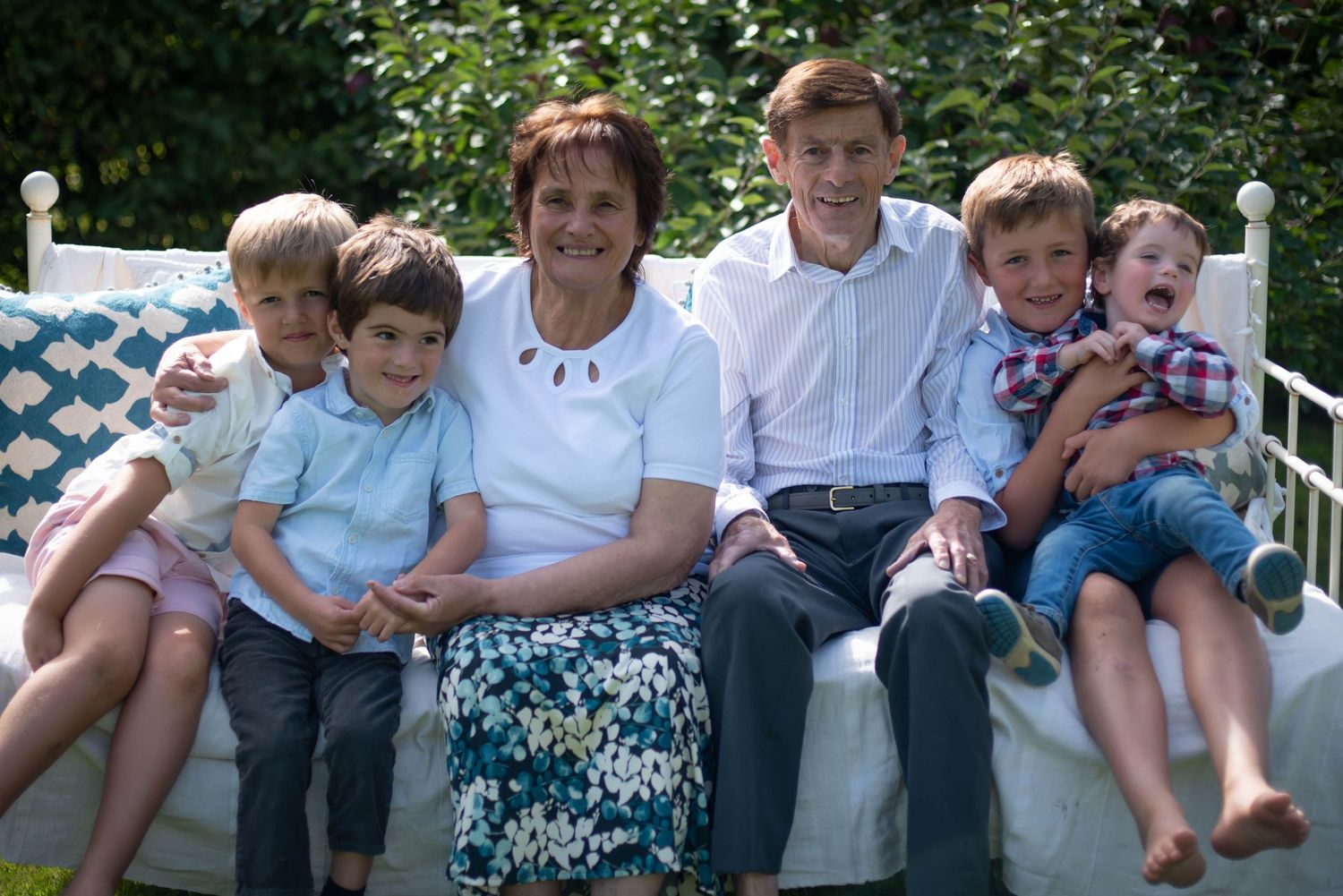 Grandparents seated on a garden bench surrounded by four young grandchildren smiling.