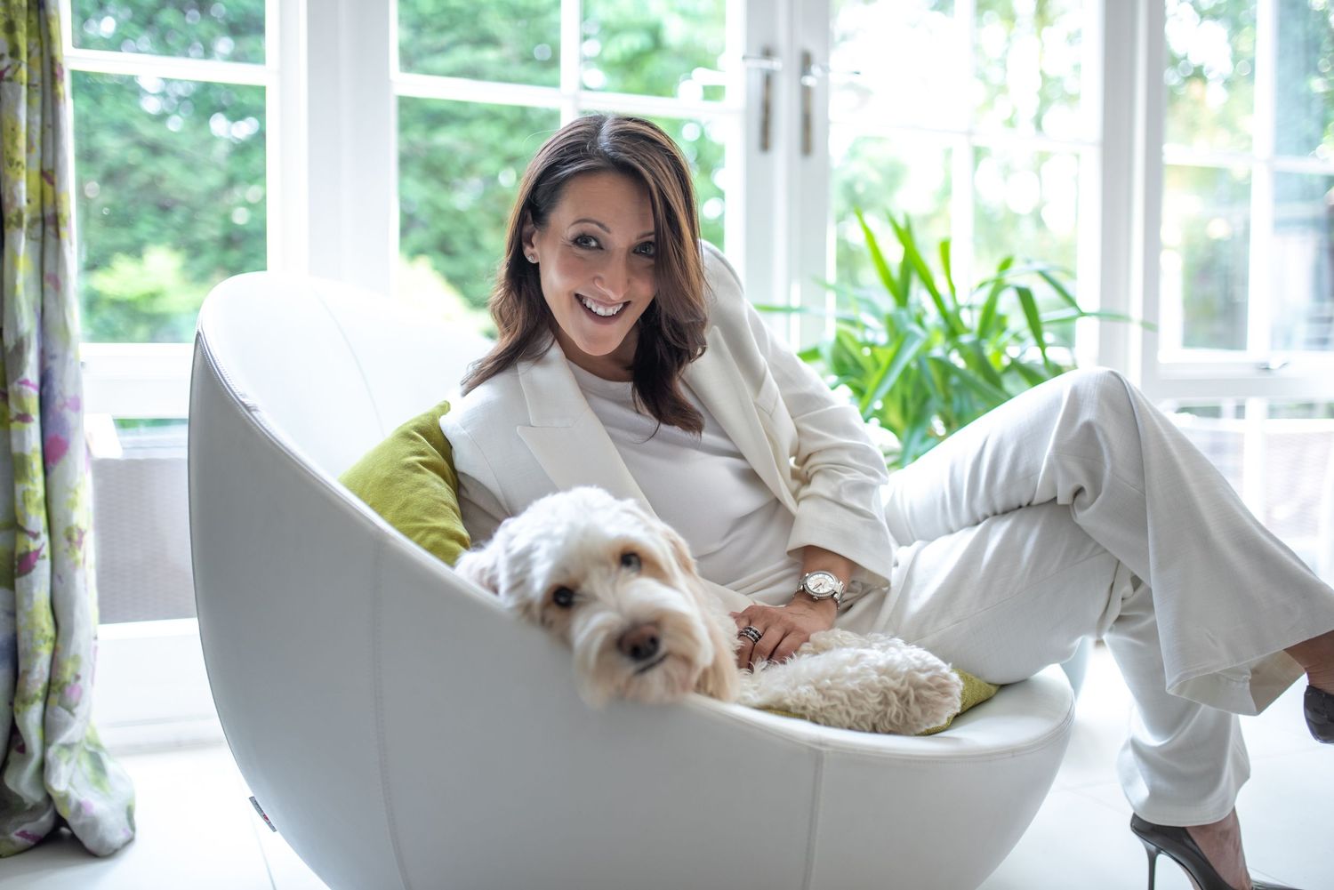 Smiling woman relaxes in a modern white chair with a fluffy dog in a bright sunlit room. Sara Davison, Divorce coach