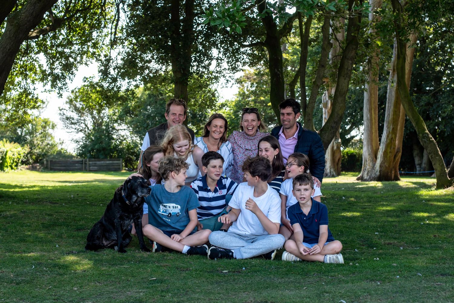 Extended family group photo on grass with large trees, adults standing behind children and black dog seated in front.