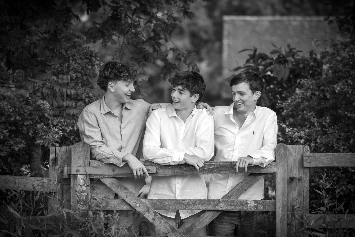 Black and white portrait of three young men laughing together leaning on a wooden gate.