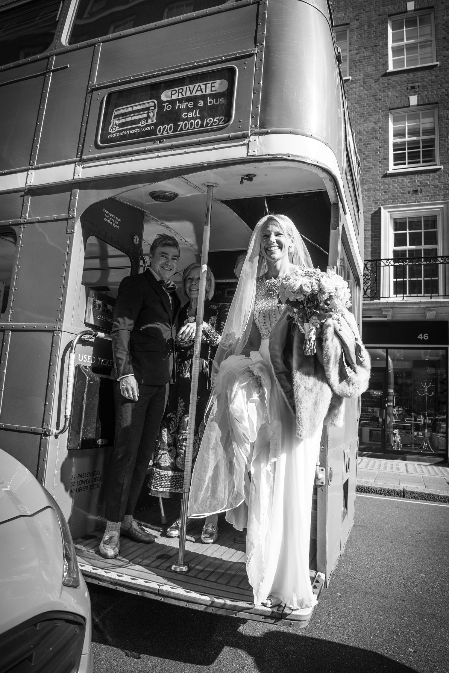Bride holding bouquet and groom standing at rear of private hire vintage red double-decker bus.
London Wedding, 34 Mayfair.