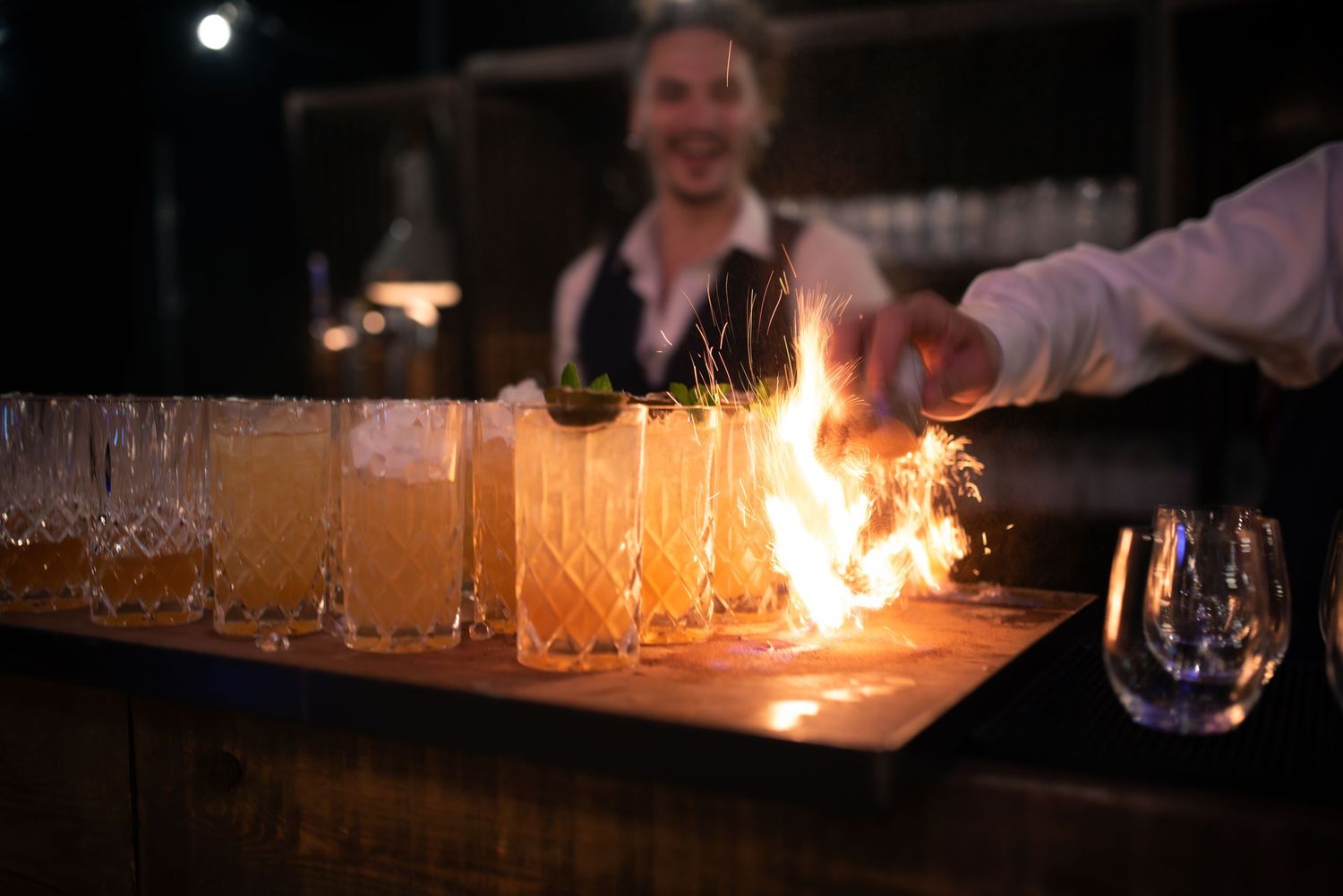 Bartender dramatically flaming cocktails lined up on wooden bar in dark atmospheric setting.
