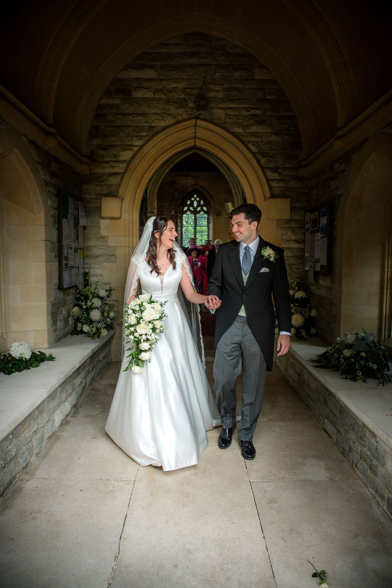 Bride and groom walking through a stone church porch, smiling at each other, bride carrying white flowers. St Saviours Church, Brockenhurst