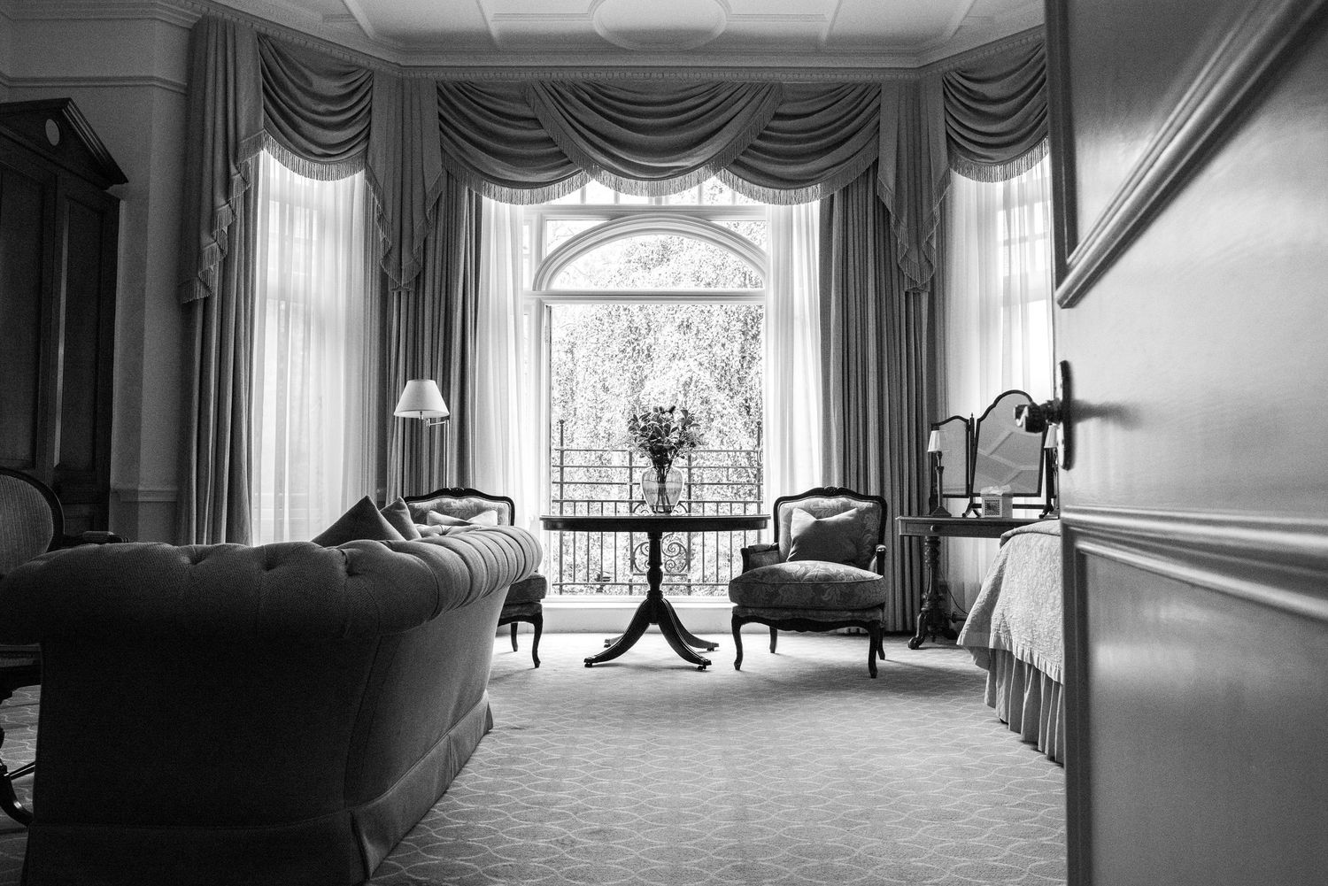 Black and white photo of a classic sitting room with tufted sofa and elegant bay window drapes. Draycott Hotel, London