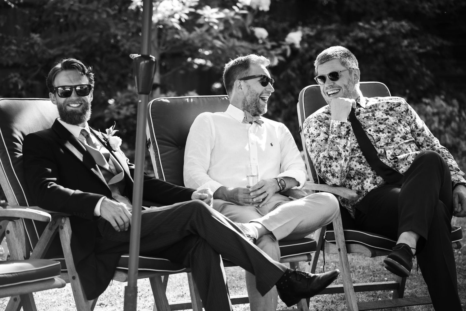 Three stylish groomsmen in sunglasses relax on lounge chairs laughing at outdoor wedding reception.