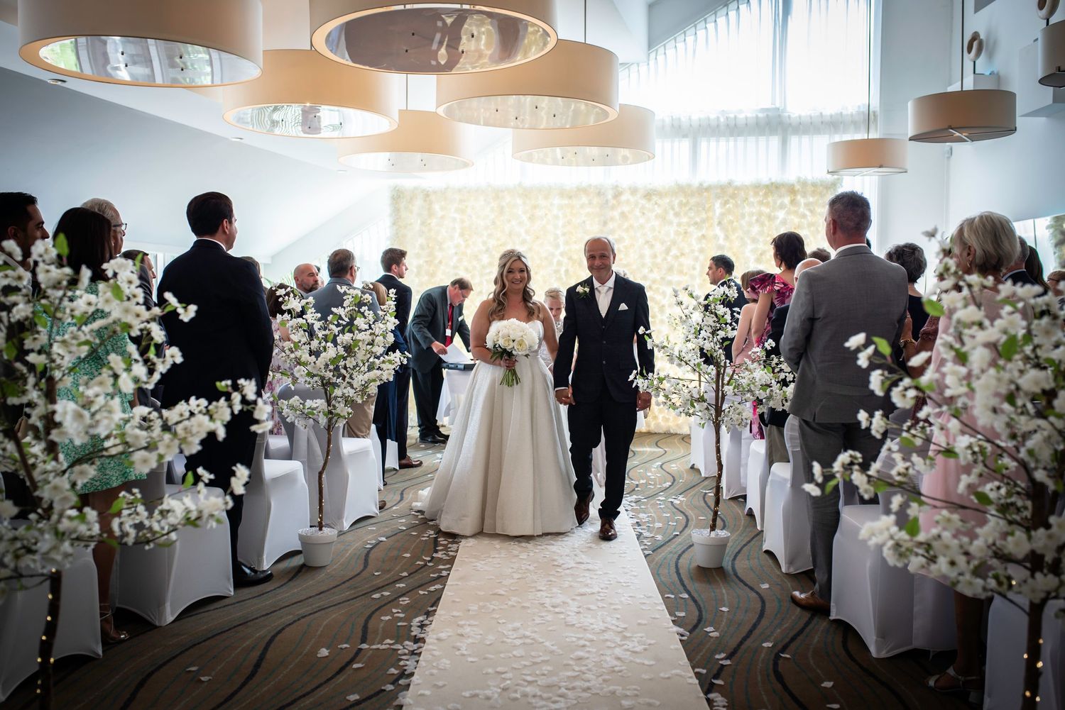 Bride and groom walking down petal-lined aisle flanked by cherry blossom trees after ceremony.