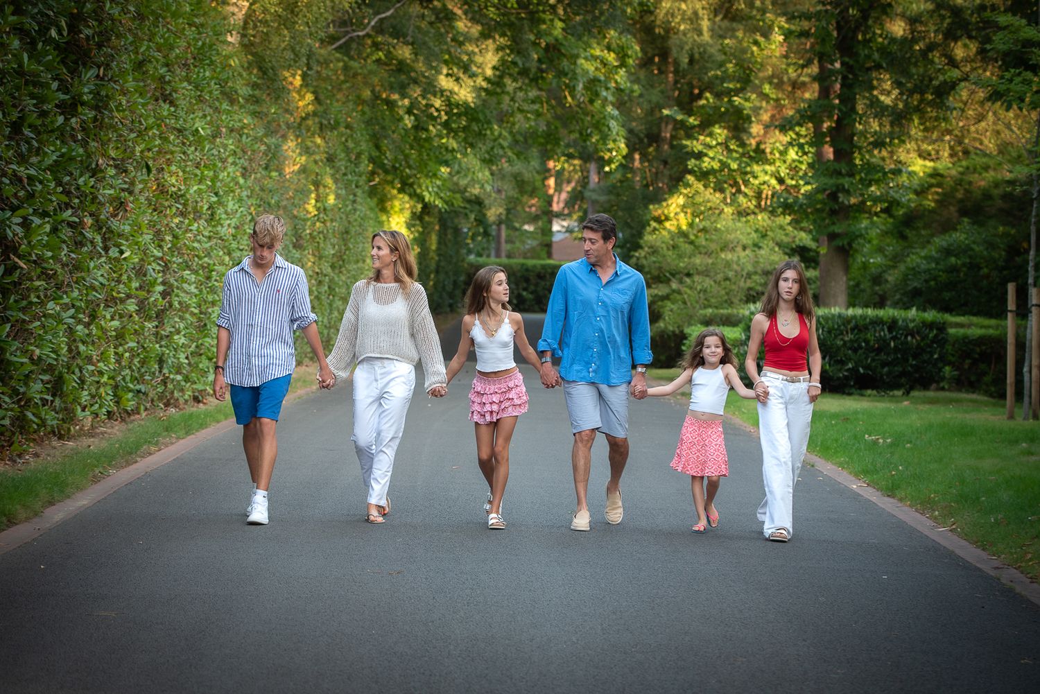 Family of five holding hands walking down sunny tree-lined path together.