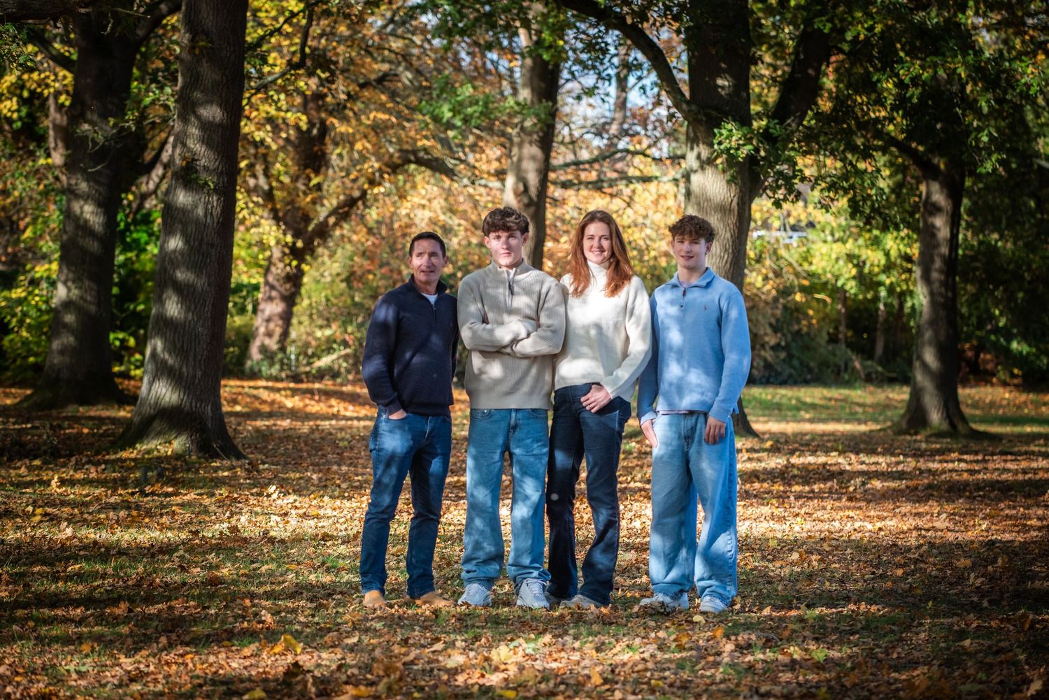 Family of four posing together in an autumn park surrounded by trees with golden fall foliage.