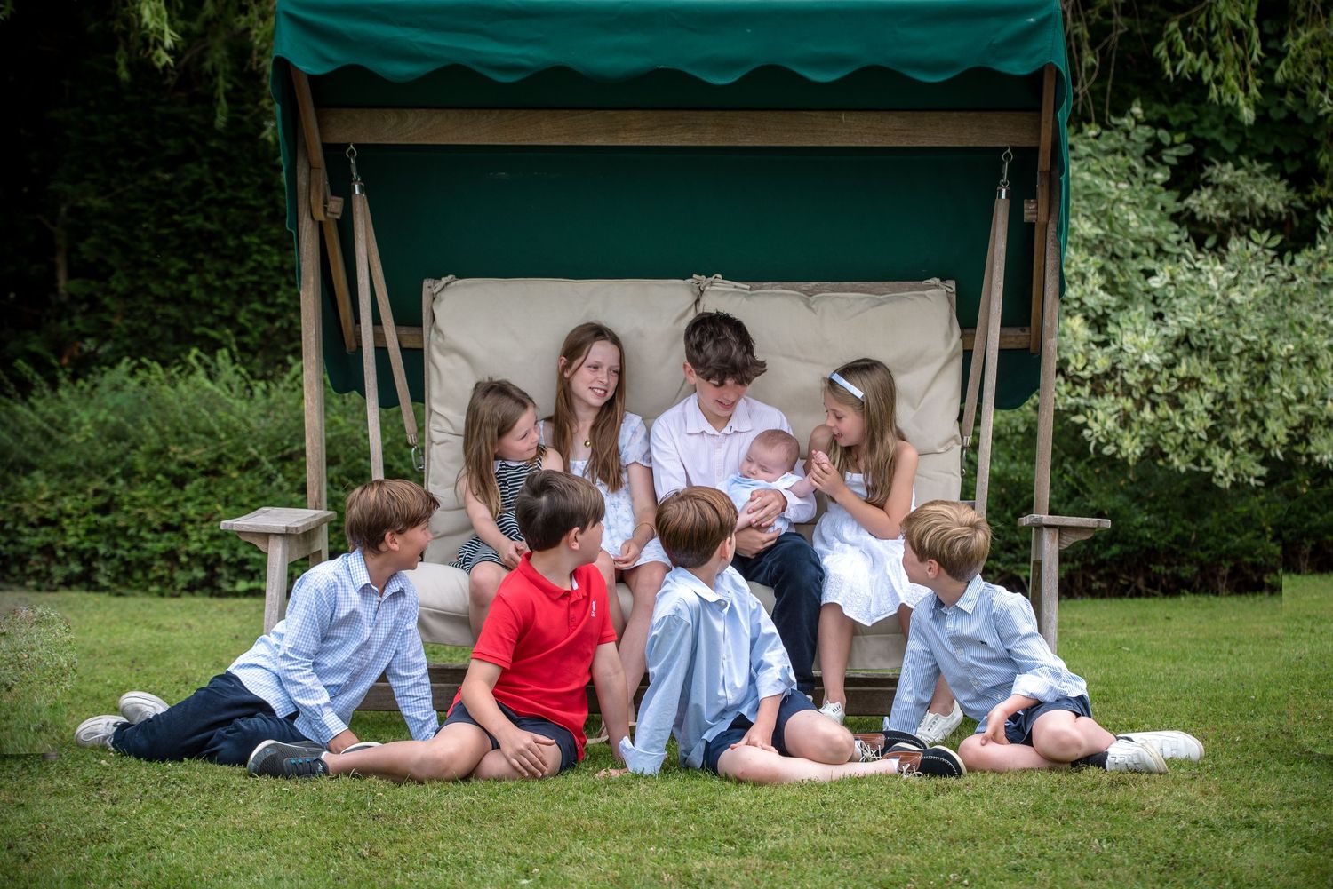 Group of children sitting together on lawn by garden swing seat in summer.