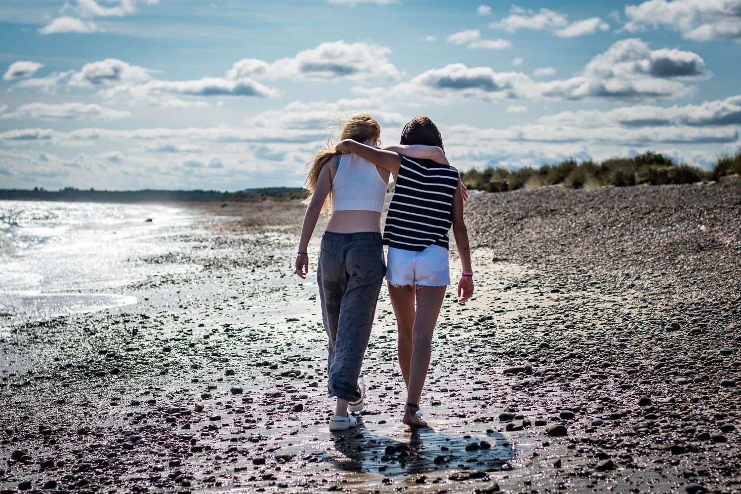 Two teenage girls walking arm in arm along a scenic coastal beach under cloudy sky.