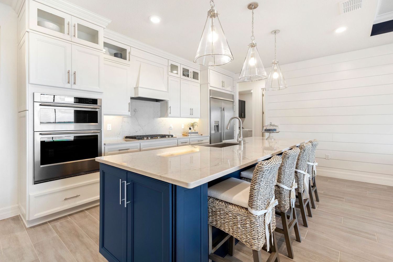 Modern kitchen with navy blue island, white cabinets, quartz countertops, pendant lights, and woven bar stools on light hardwood floors.