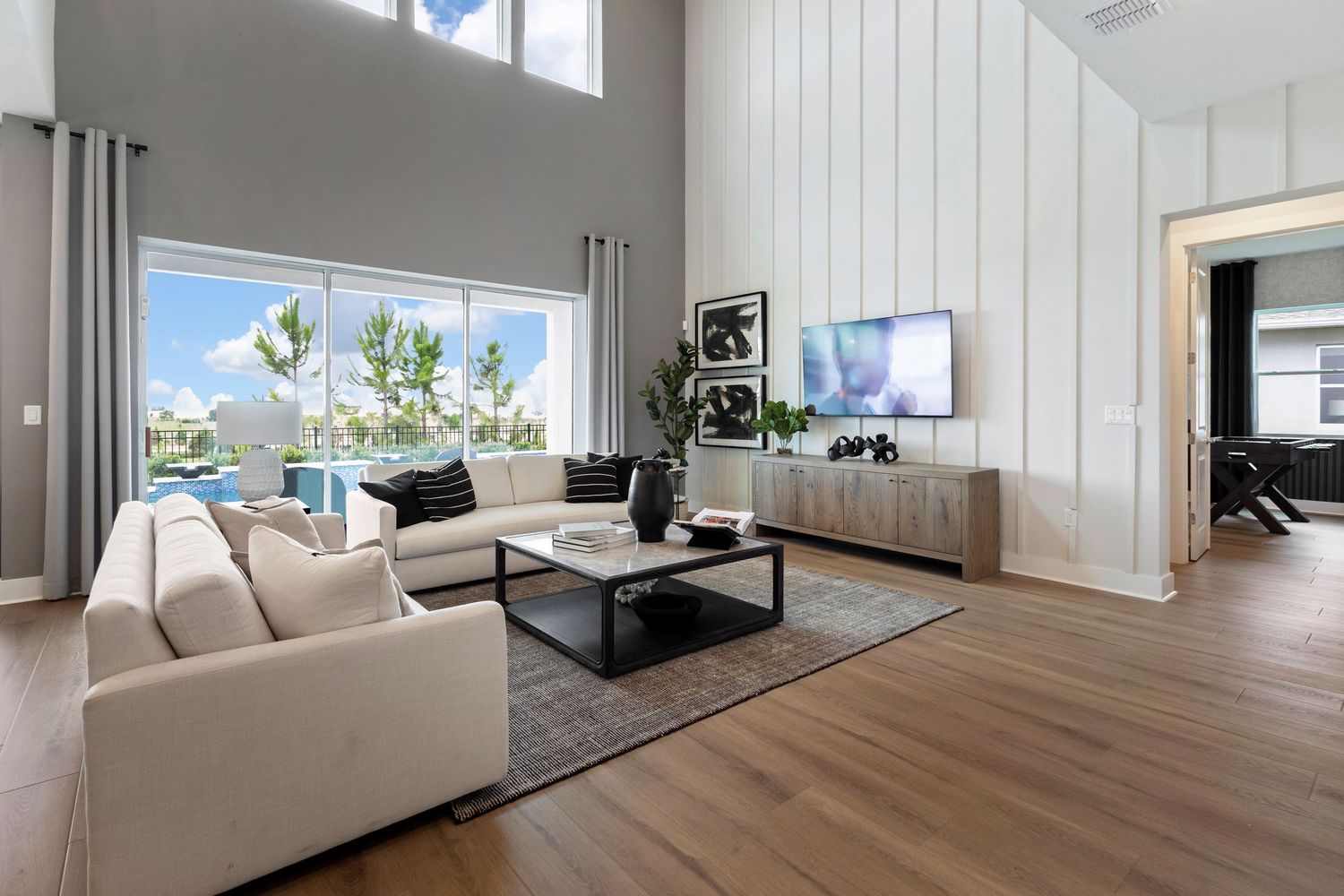 Modern living room with high ceilings, white shiplap walls, beige sectional sofa, wood floors, and large windows with pool view.