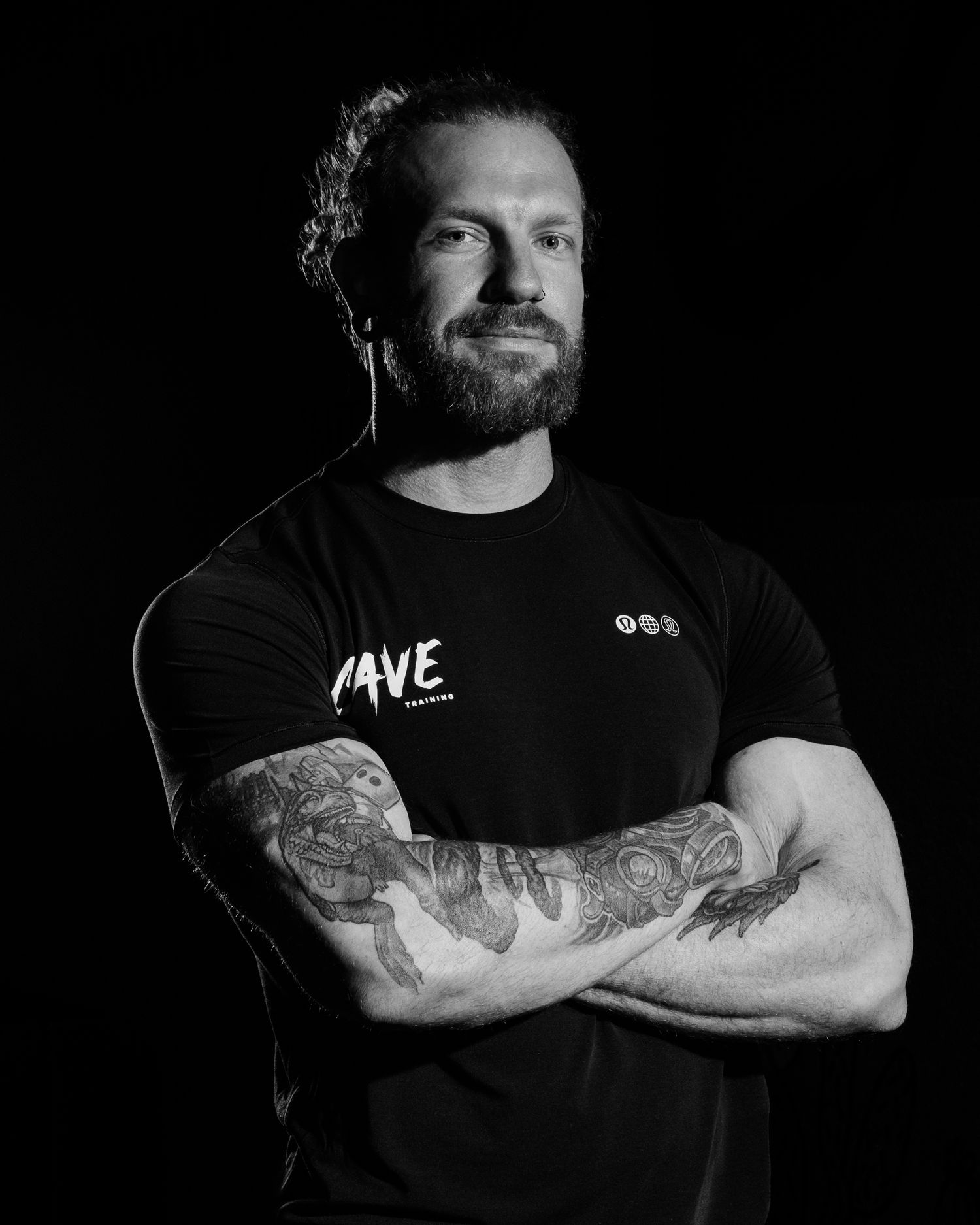 Black and white portrait of a tattooed bearded man in a black Cave Training shirt, arms crossed.